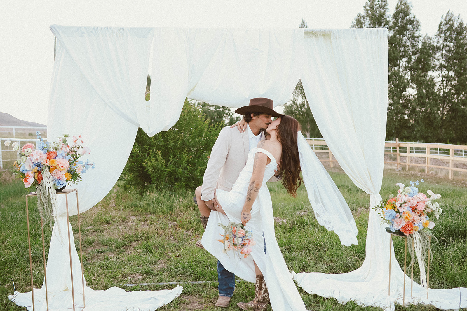 Two women in white dresses and hats embrace under a white fabric canopy with floral arrangements on either side, outdoors.
