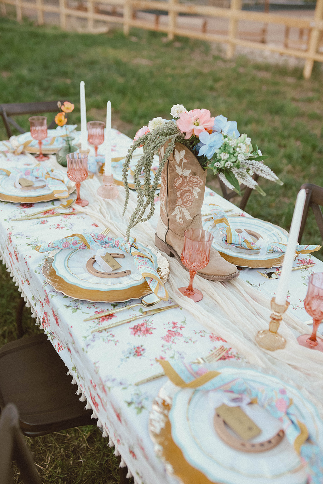 Outdoor table set with floral centerpiece, candles, plates, and decorated cookies, on a grassy area with a wooden fence in background.