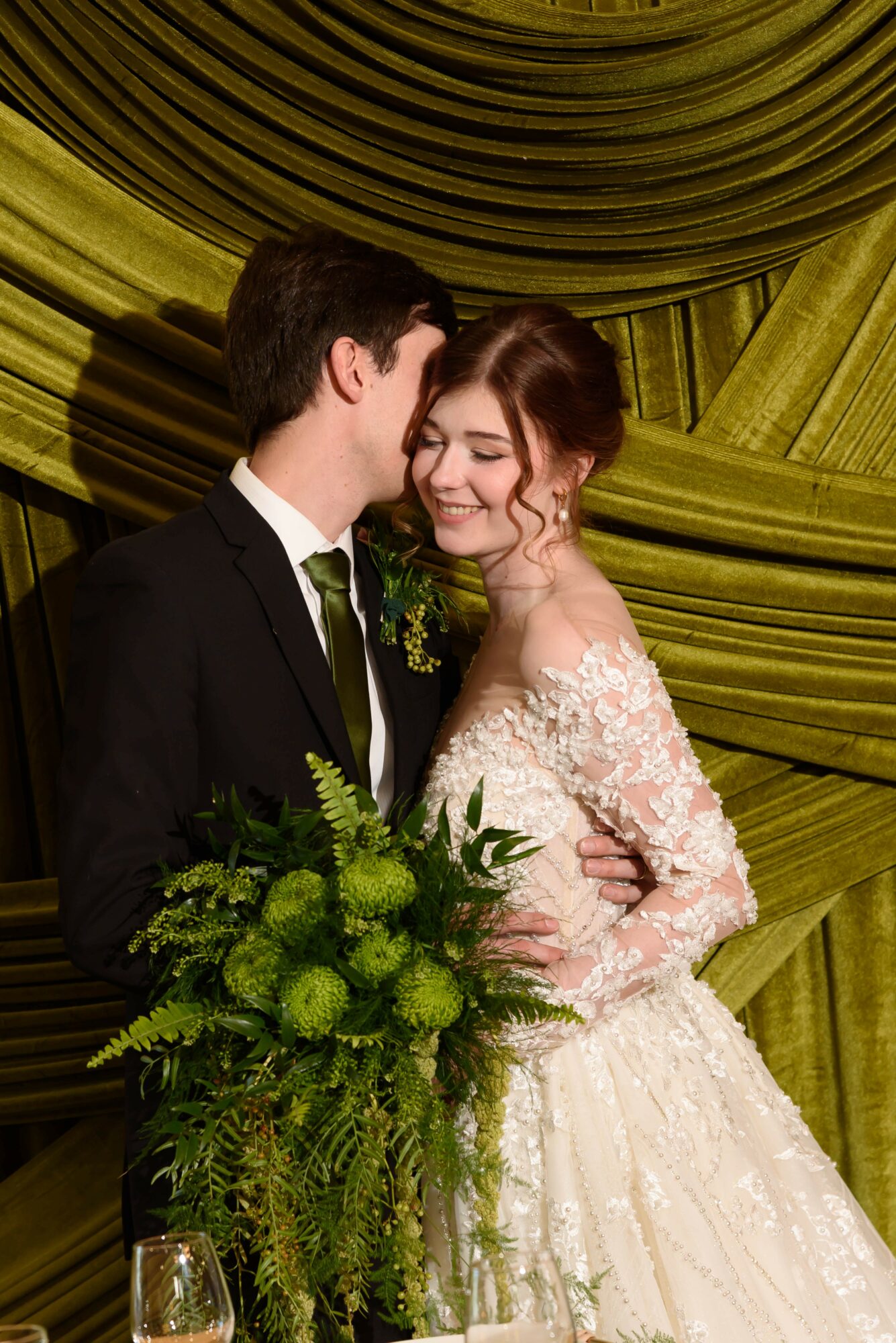 A bride and groom smile and embrace, with the bride holding a large green floral bouquet, against a green draped background.