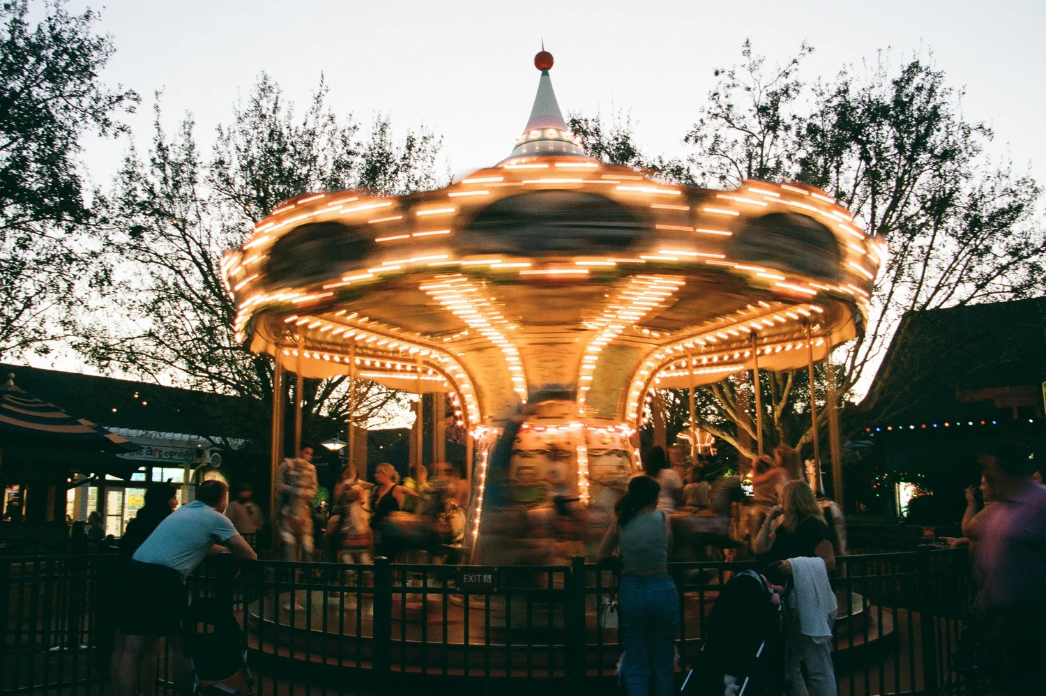 People riding a carousel with lights, trees, and sky in the background, some standing and watching, during evening.