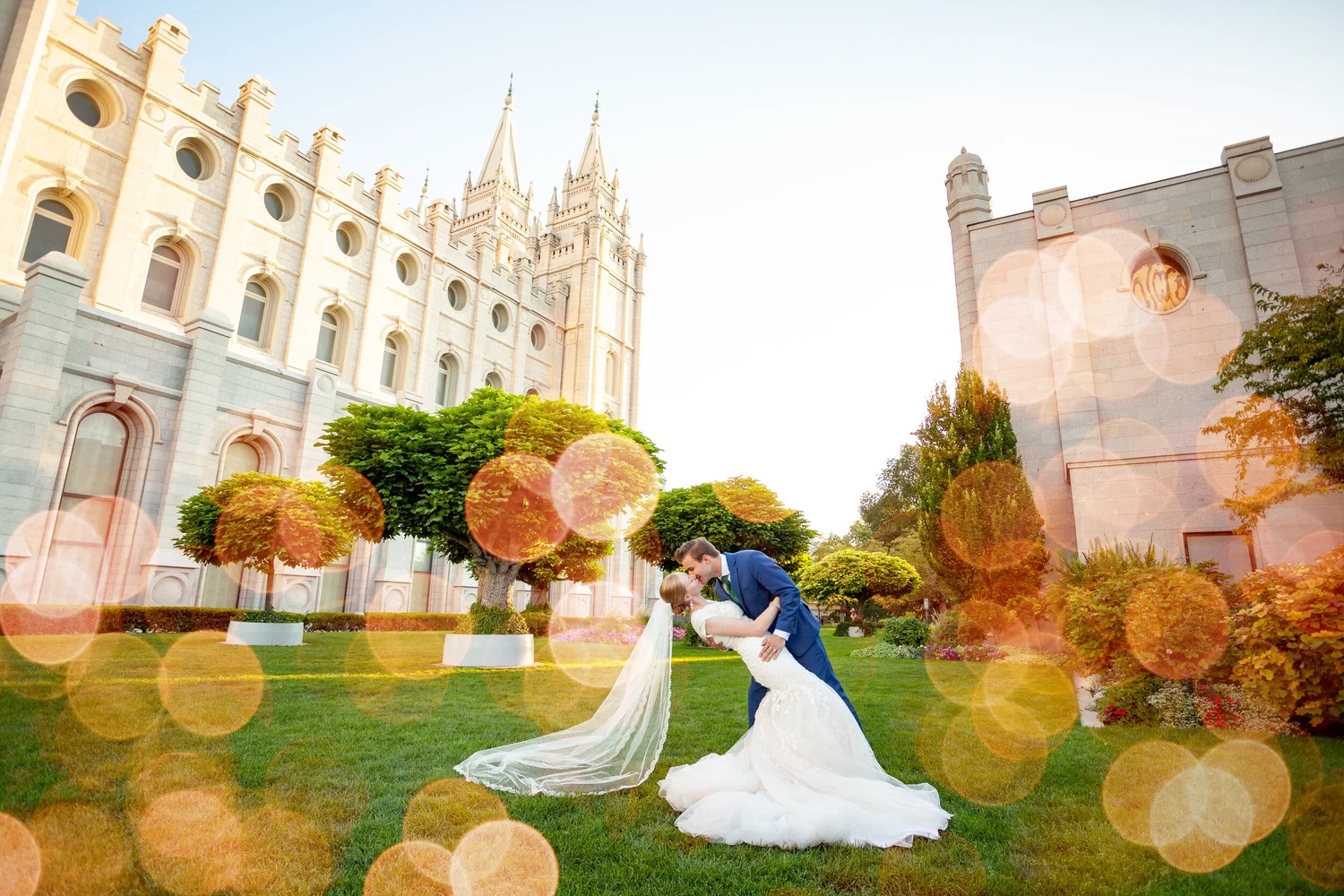 Bride and groom kissing on a lawn with historic buildings and trees in the background, sunlight creating lens flare effects.
