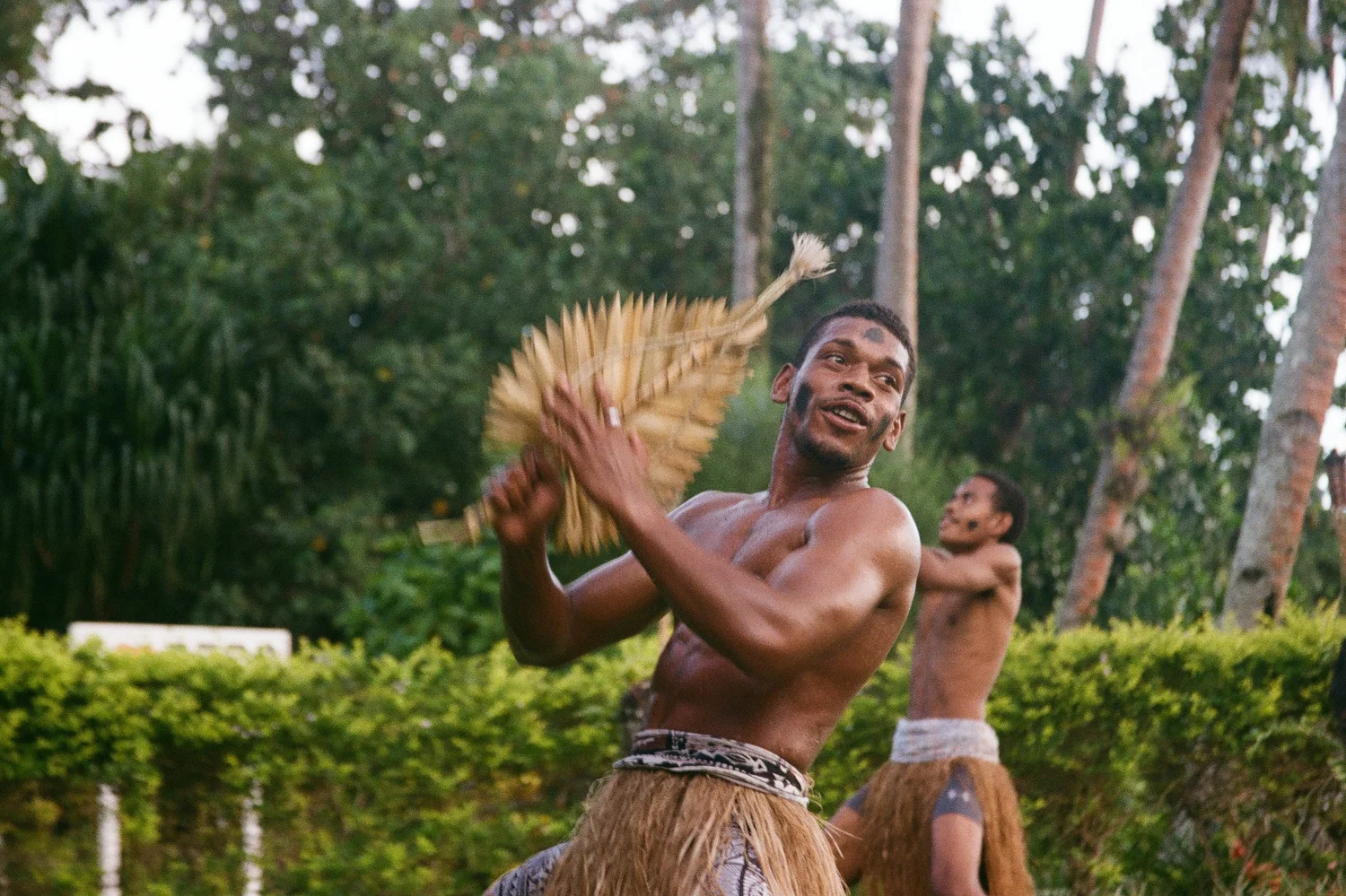 Two shirtless men outdoors, one clapping with a fan, surrounded by trees and greenery.
