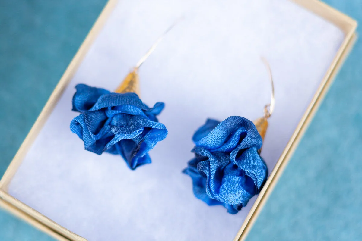 Pair of blue fabric flower earrings with gold hooks on a white background, in a box.