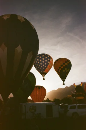 Three hot air balloons in the sky during sunset, with vehicles and people on the ground, clouds in the background.