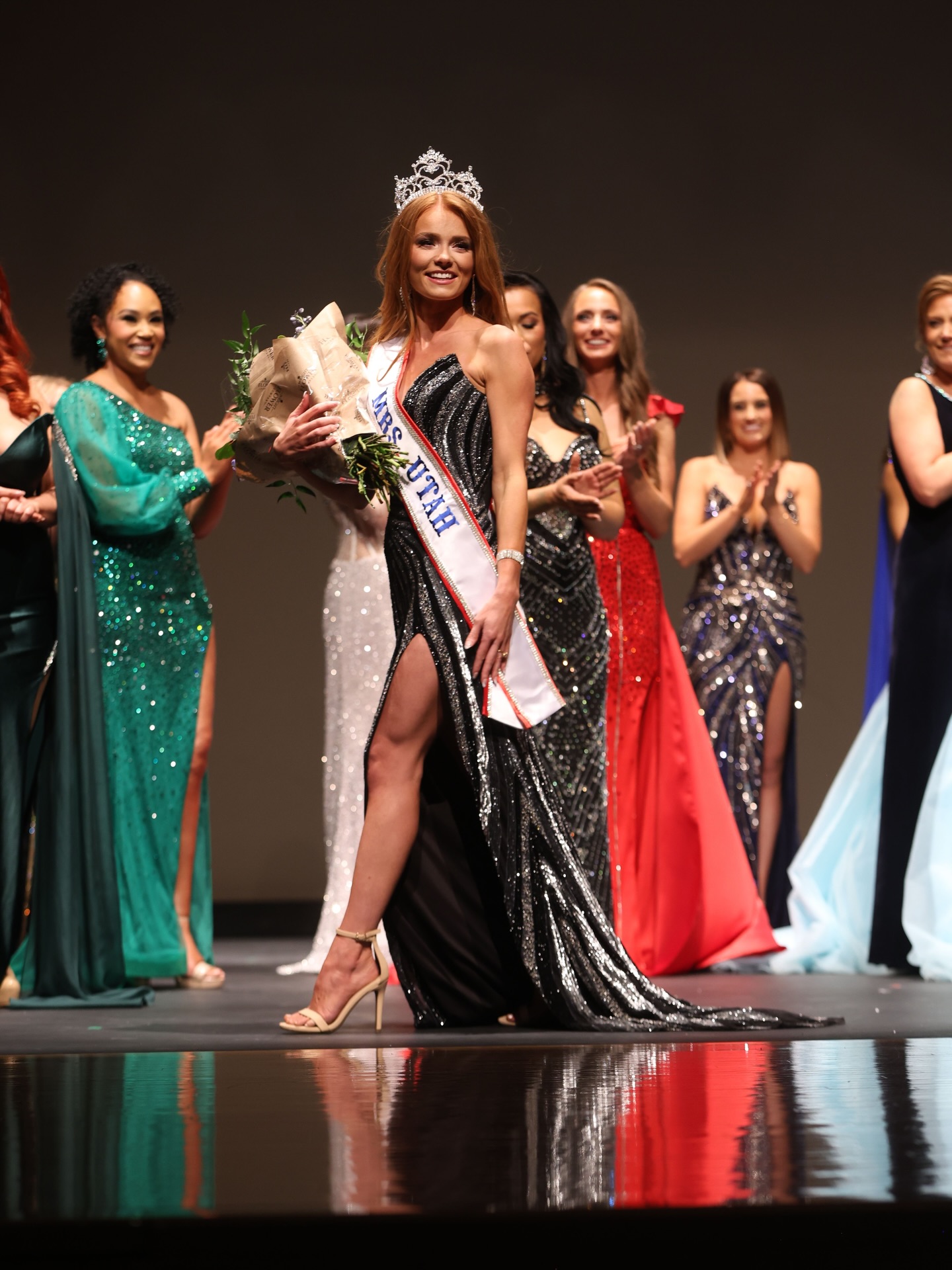 Woman wearing a crown and sash holding flowers, surrounded by women in evening gowns on stage.