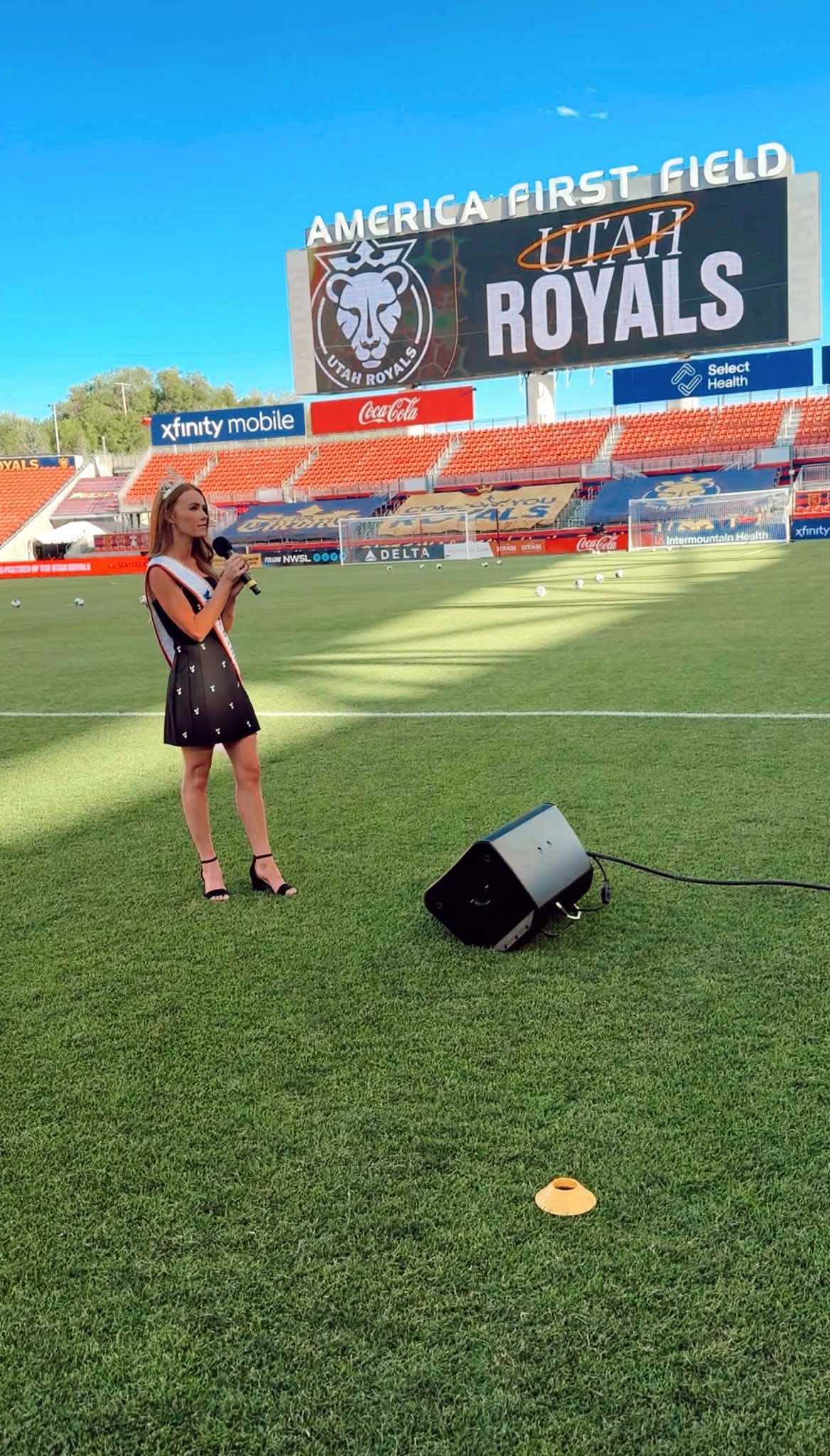Young girl singing into a microphone on a sports field with stadium seating and signs in the background.