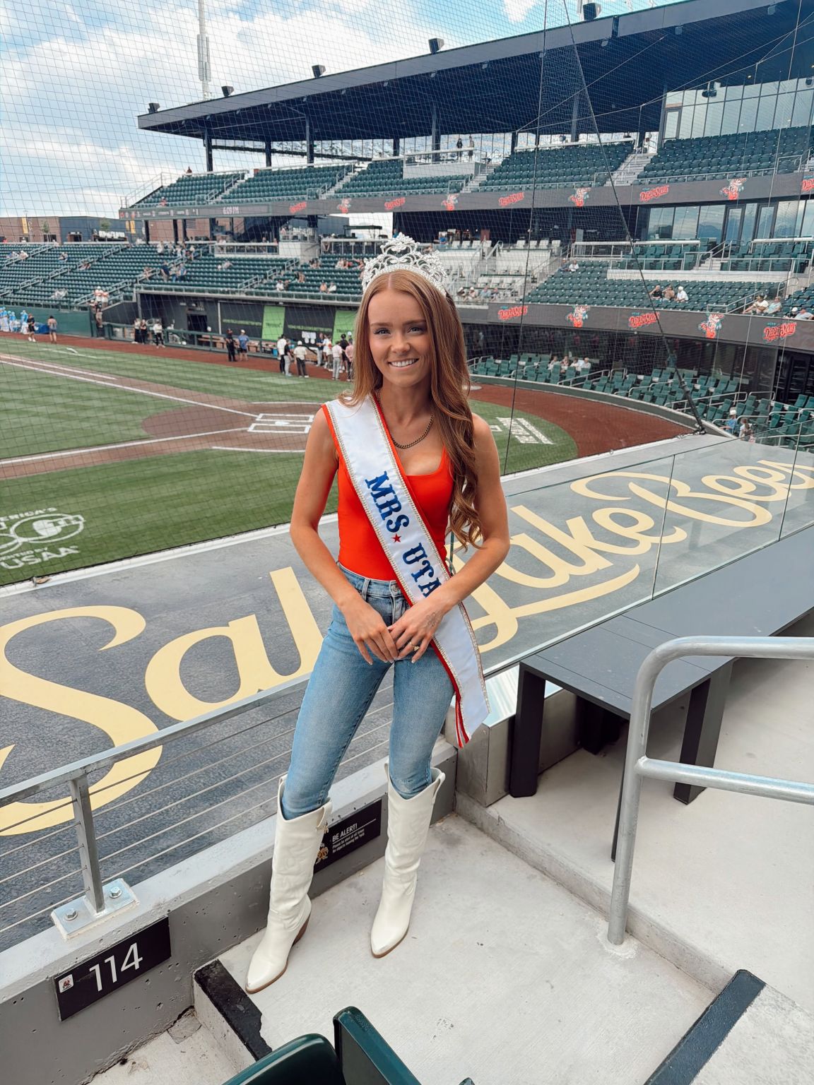 Young woman in sash and jeans standing in a baseball stadium, smiling, with empty seats and field in background.