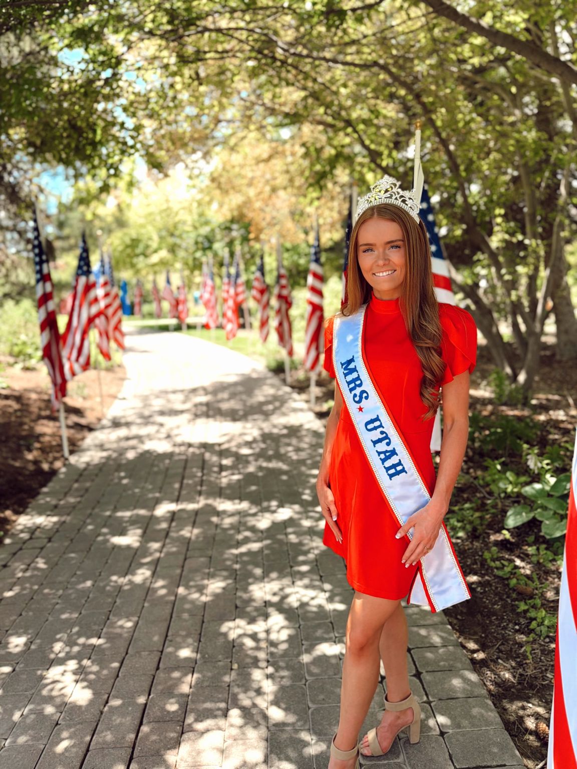 Young girl in a red dress with a crown and sash standing on a path lined with American flags, trees in background.