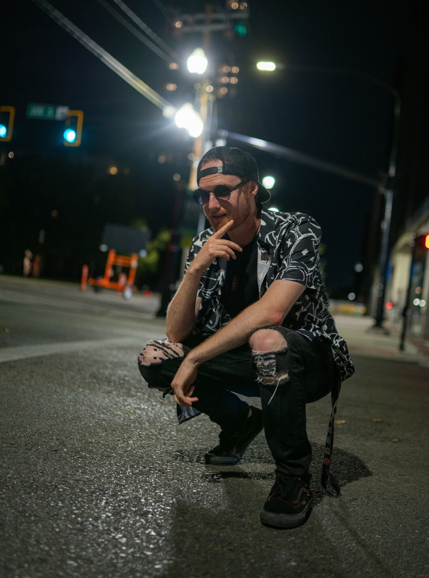 Person crouching on street at night, wearing sunglasses, patterned shirt, ripped jeans, with streetlights and traffic signals in background.