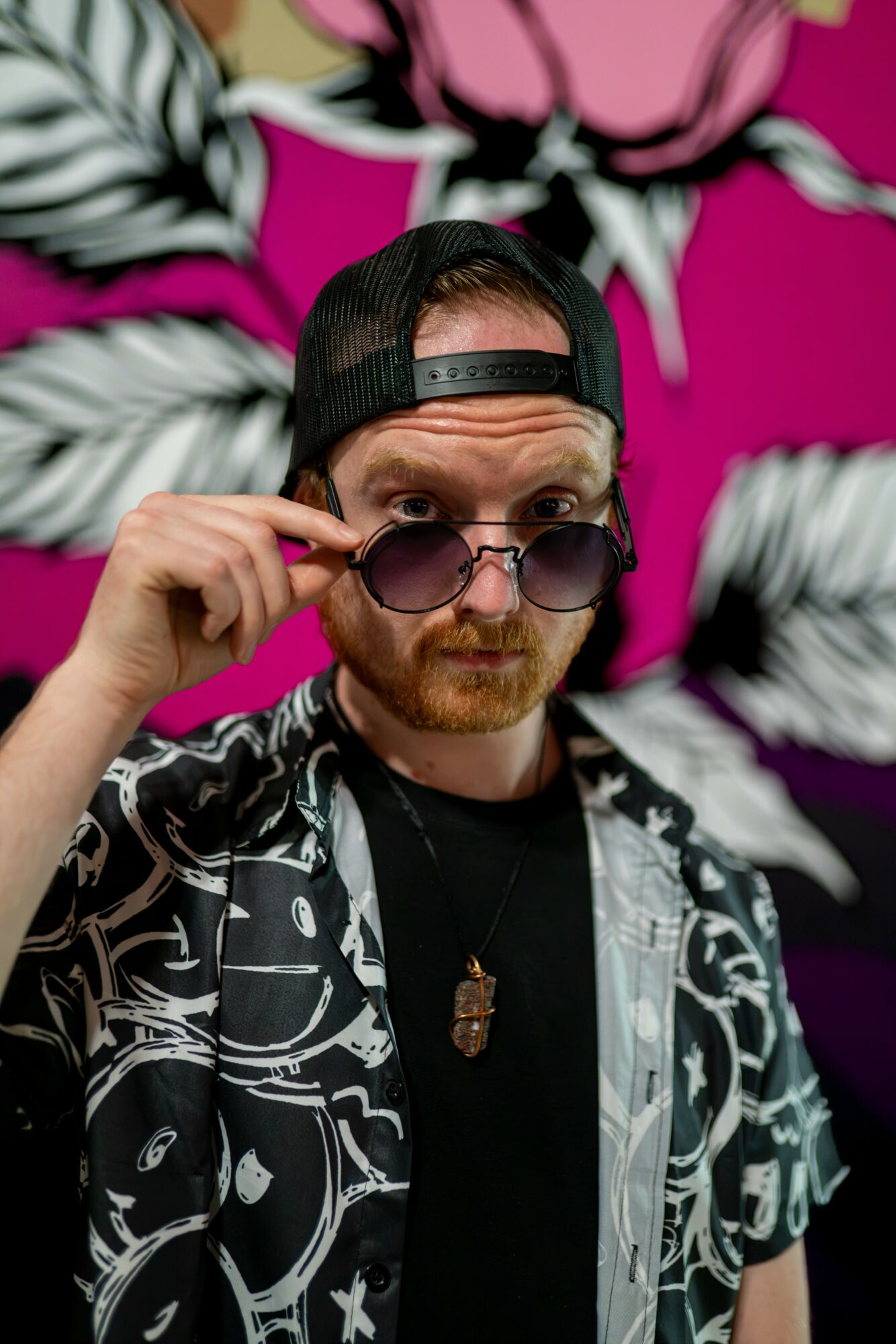 Man with sunglasses and a cap adjusting his glasses, standing in front of a colorful floral background.