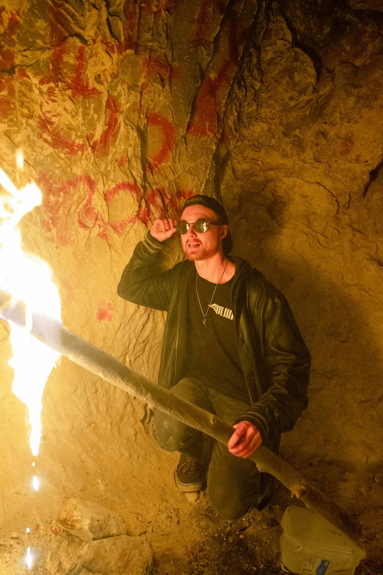 Person wearing sunglasses and a black jacket holding a large fire hose inside a cave with graffiti-covered walls.
