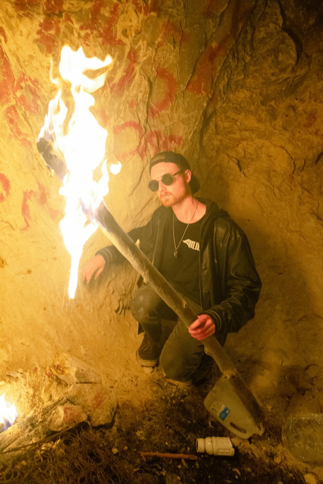 Person holding a flaming torch inside a cave, wearing sunglasses and a black jacket, kneeling on the ground.