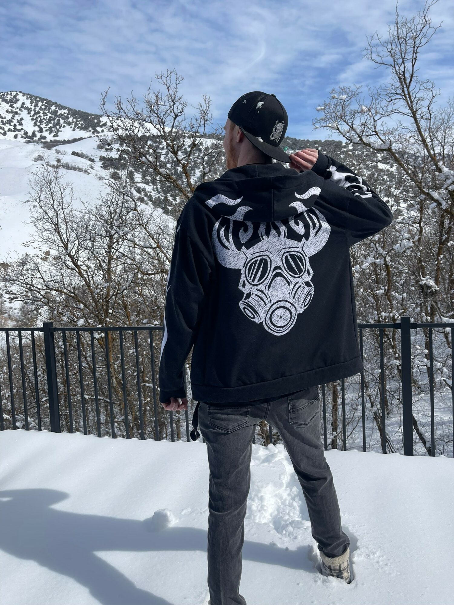 Person standing outdoors in snowy landscape, wearing a black hoodie with a gas mask graphic, facing away from camera.