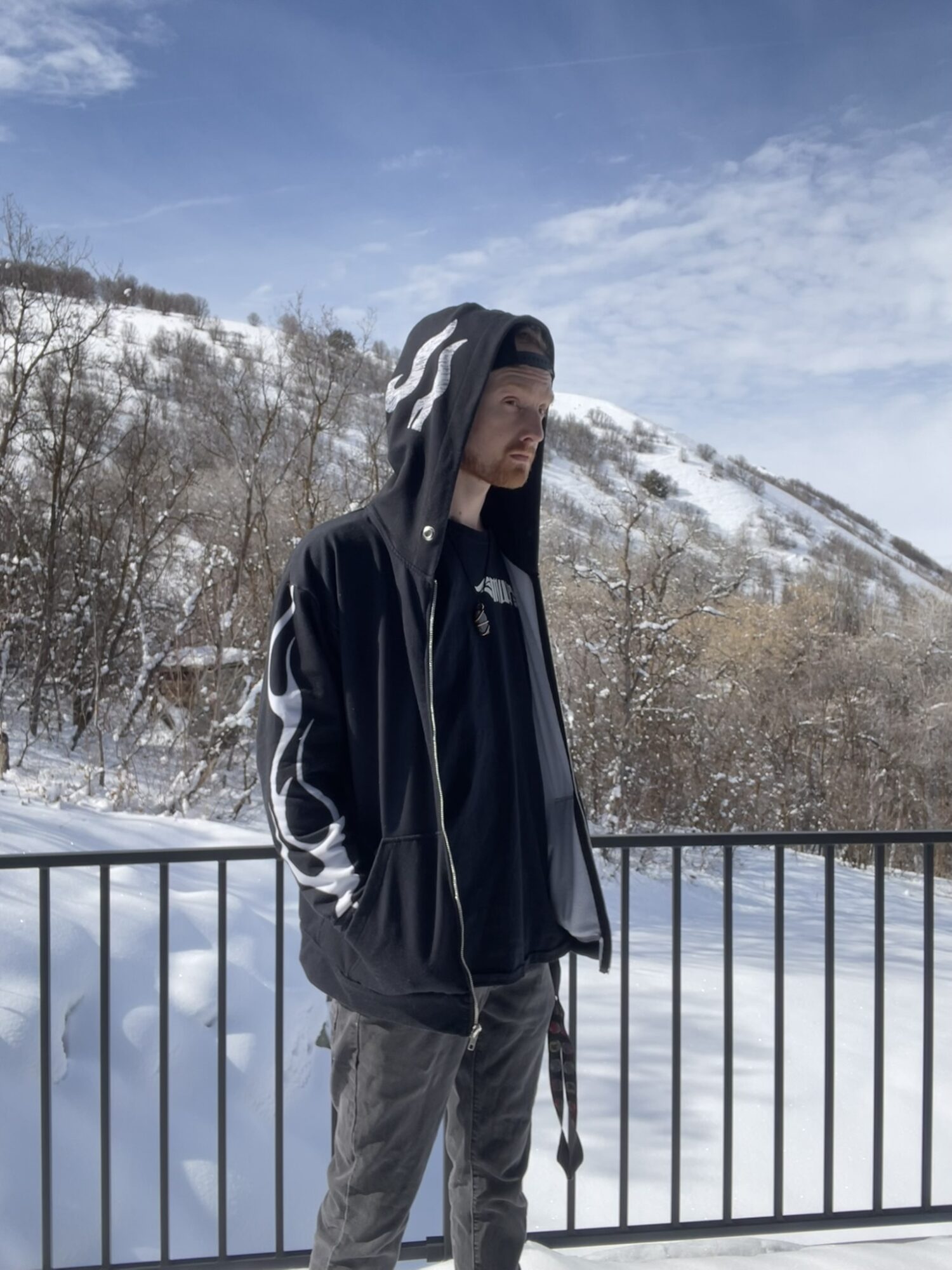 Young person wearing a black hoodie and jacket stands outdoors with snow-covered mountains and trees in background.