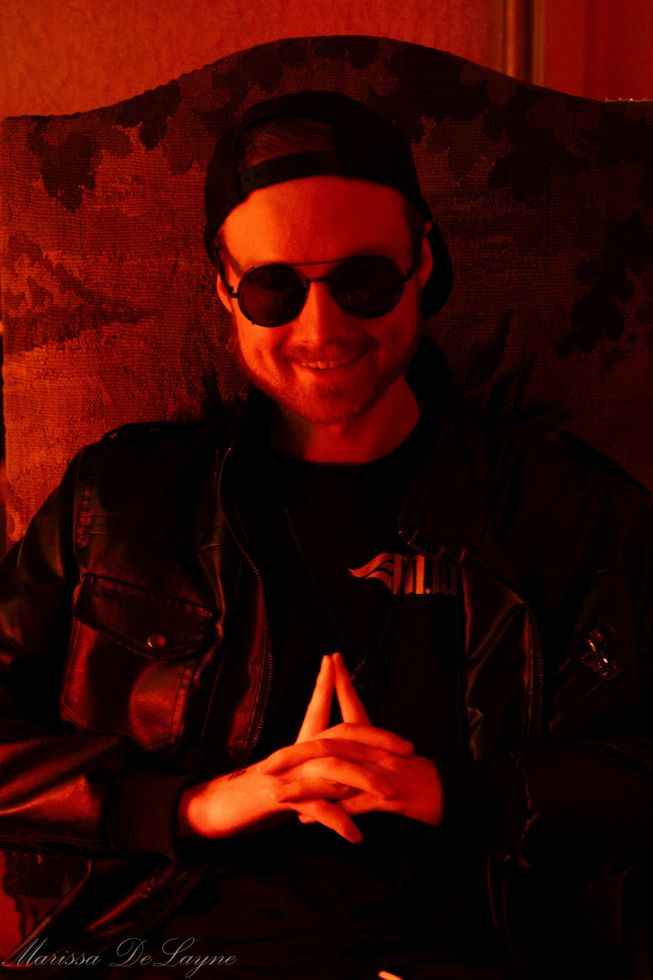 Person wearing sunglasses and a cap, smiling, sitting with hands clasped, in a dimly lit room.