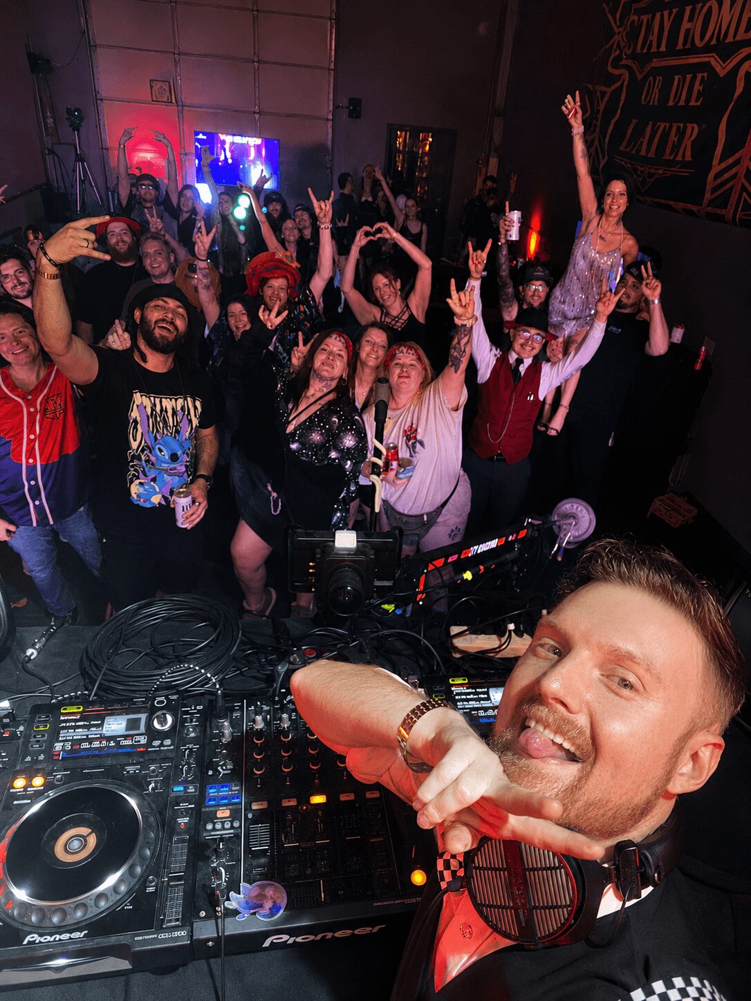 Crowd of people dancing and celebrating at a party with a DJ in the foreground, smiling and raising hands, colorful lighting.