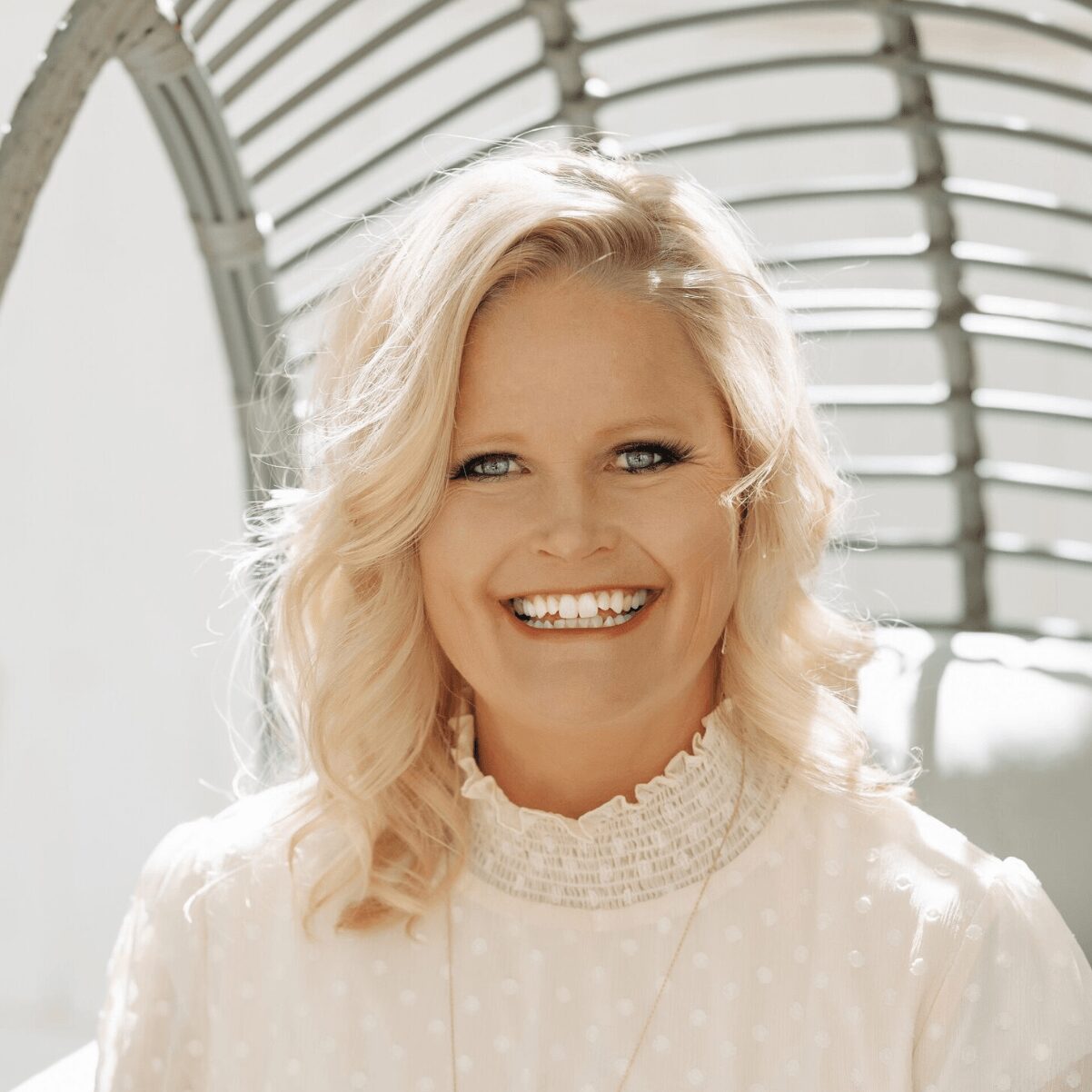Smiling woman with blonde hair in a white polka dot blouse, standing outdoors with a curved architectural background.