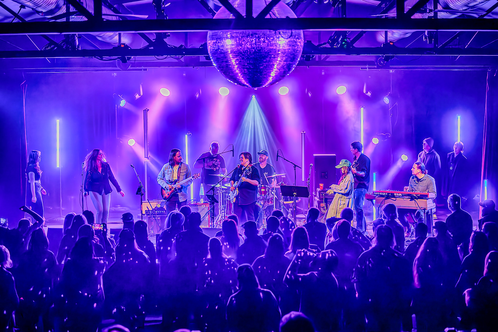 Band performs on stage with purple lighting and a large disco ball overhead, audience watching attentively.