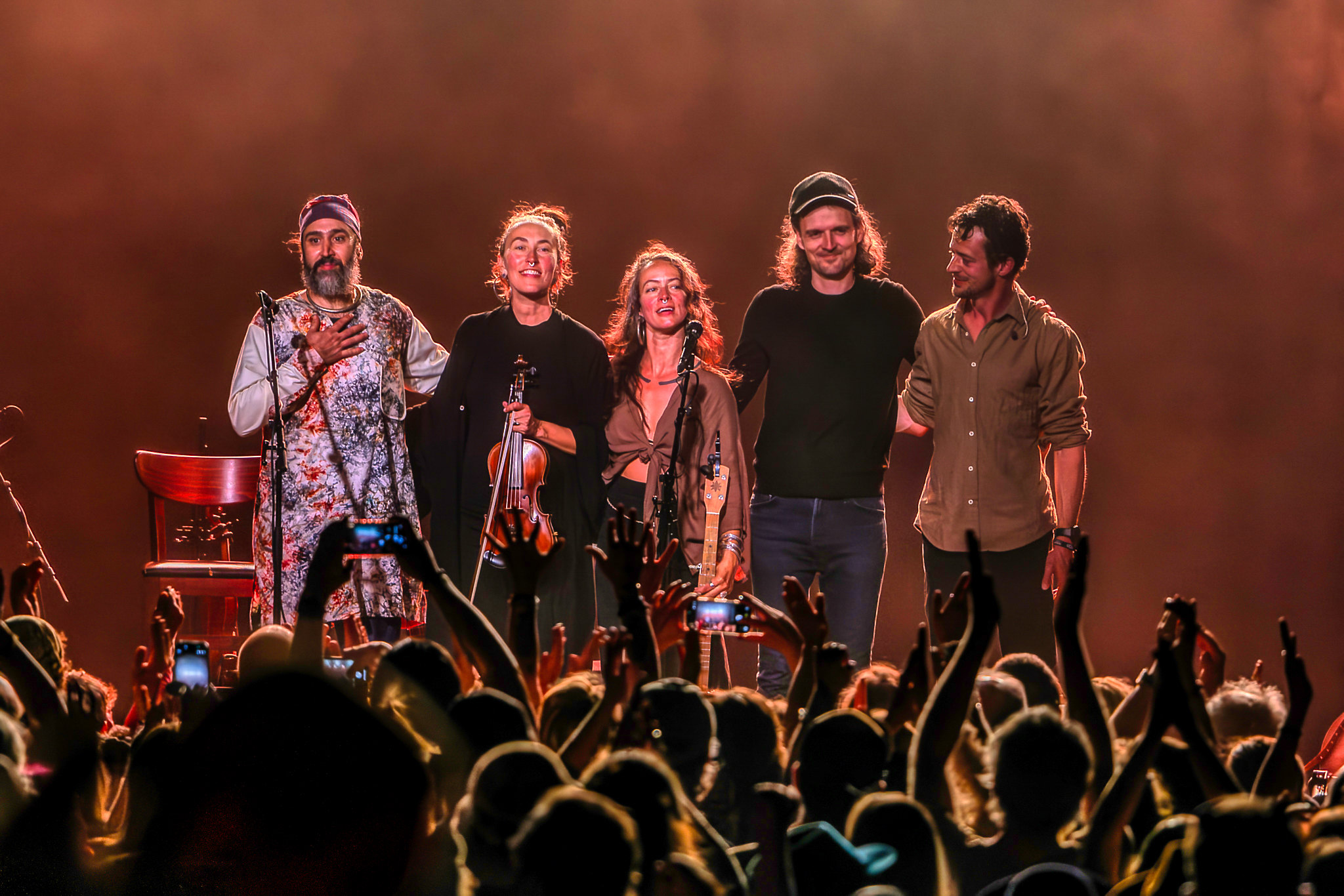 Five performers stand on stage holding microphones, with an audience in front, stage lighting in the background.