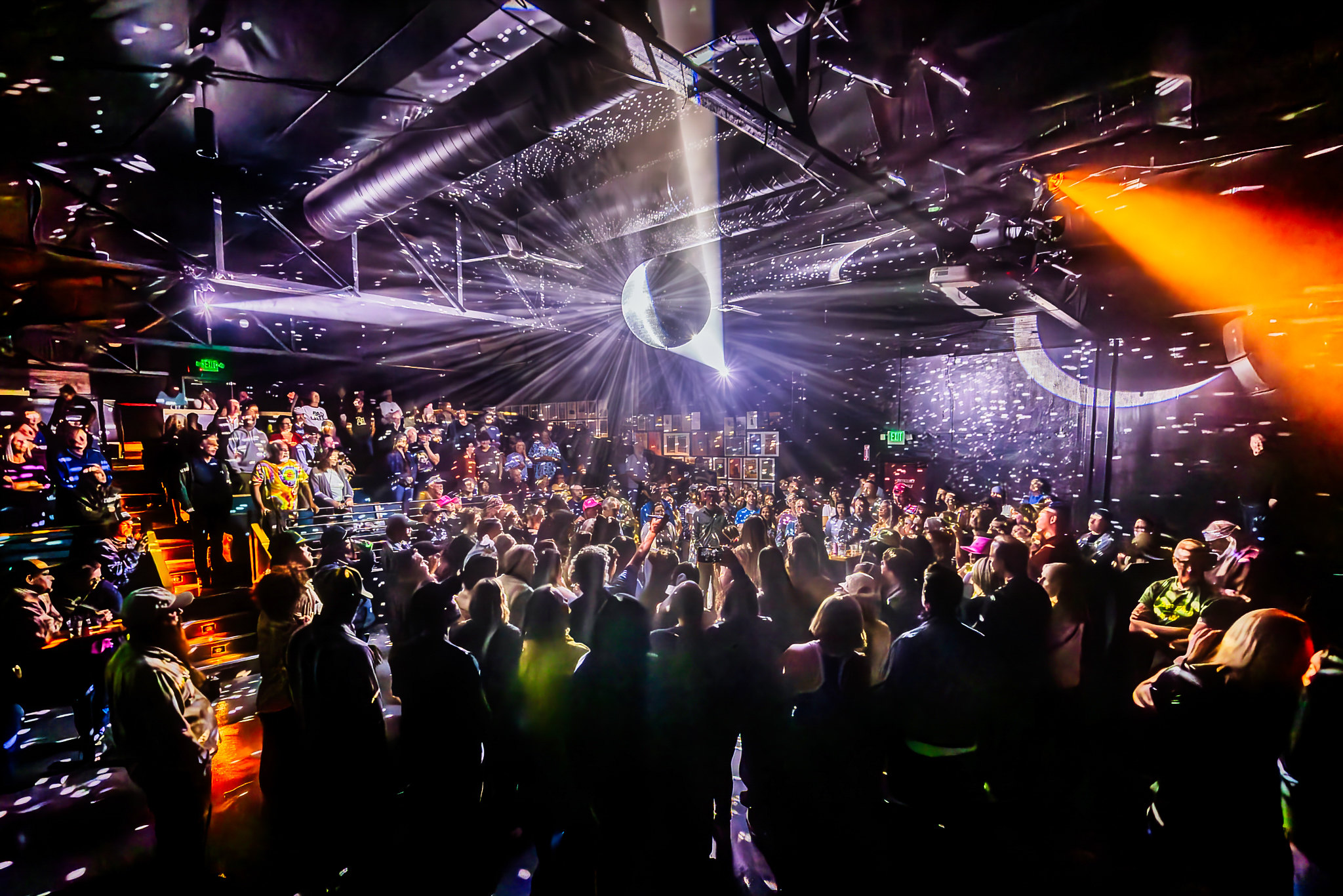 Crowd in a nightclub with colorful lights, laser beams, and a bright central light display.
