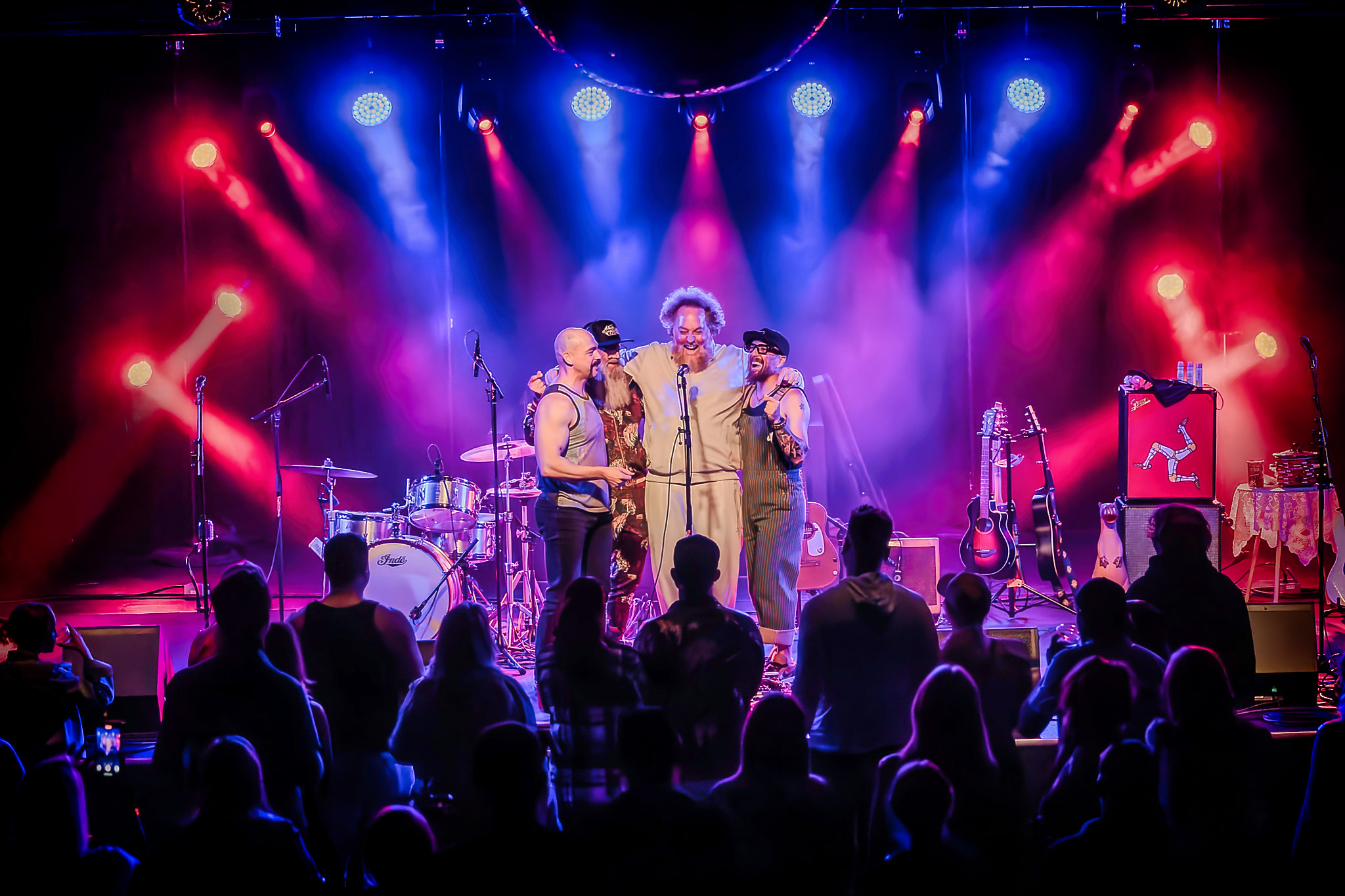 Three performers on stage with colorful lighting, audience watching, musical instruments visible, vibrant concert scene.