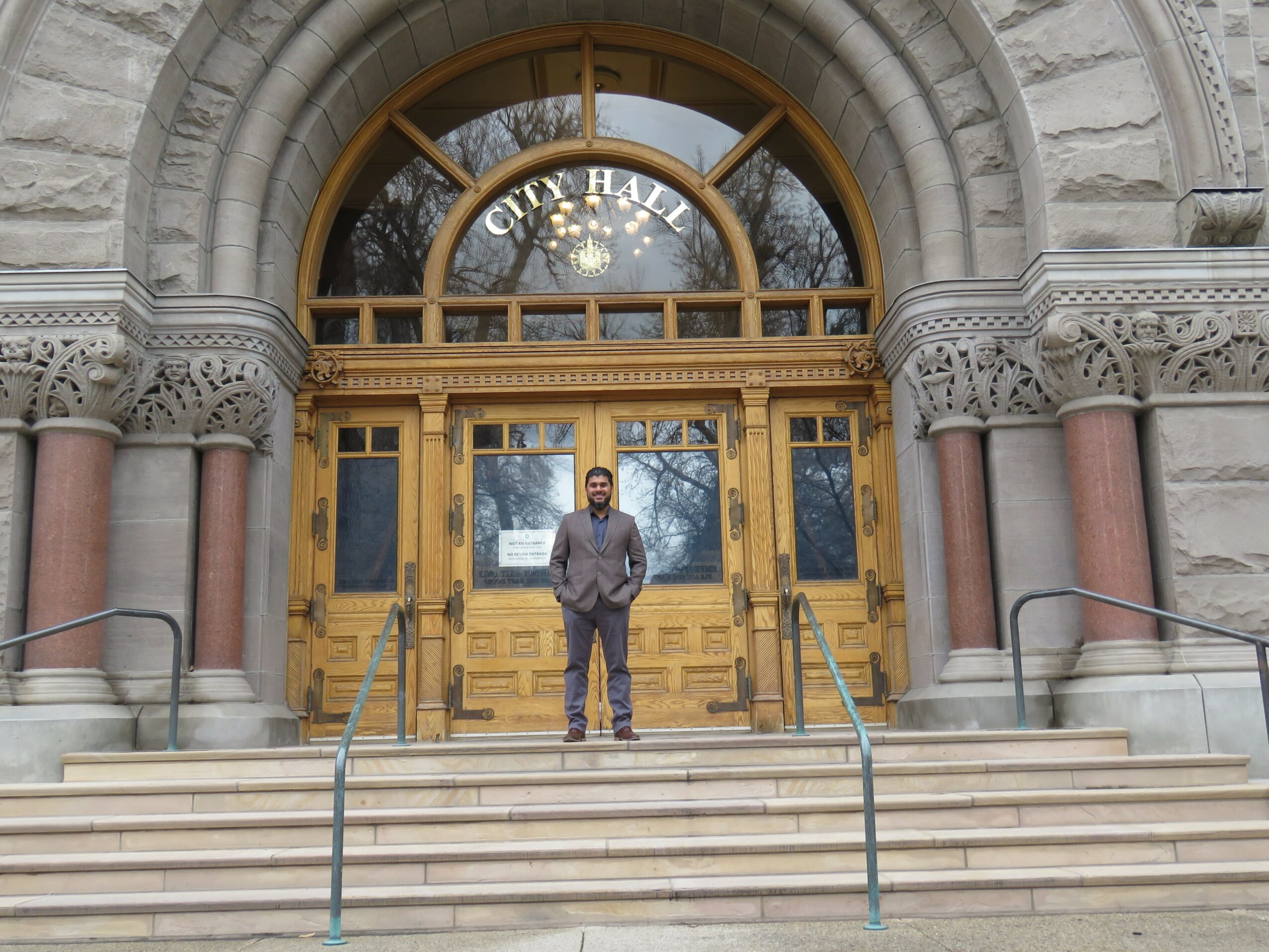 Man standing on steps in front of a large arched entrance with glass and wood doors, stone and brick architecture.