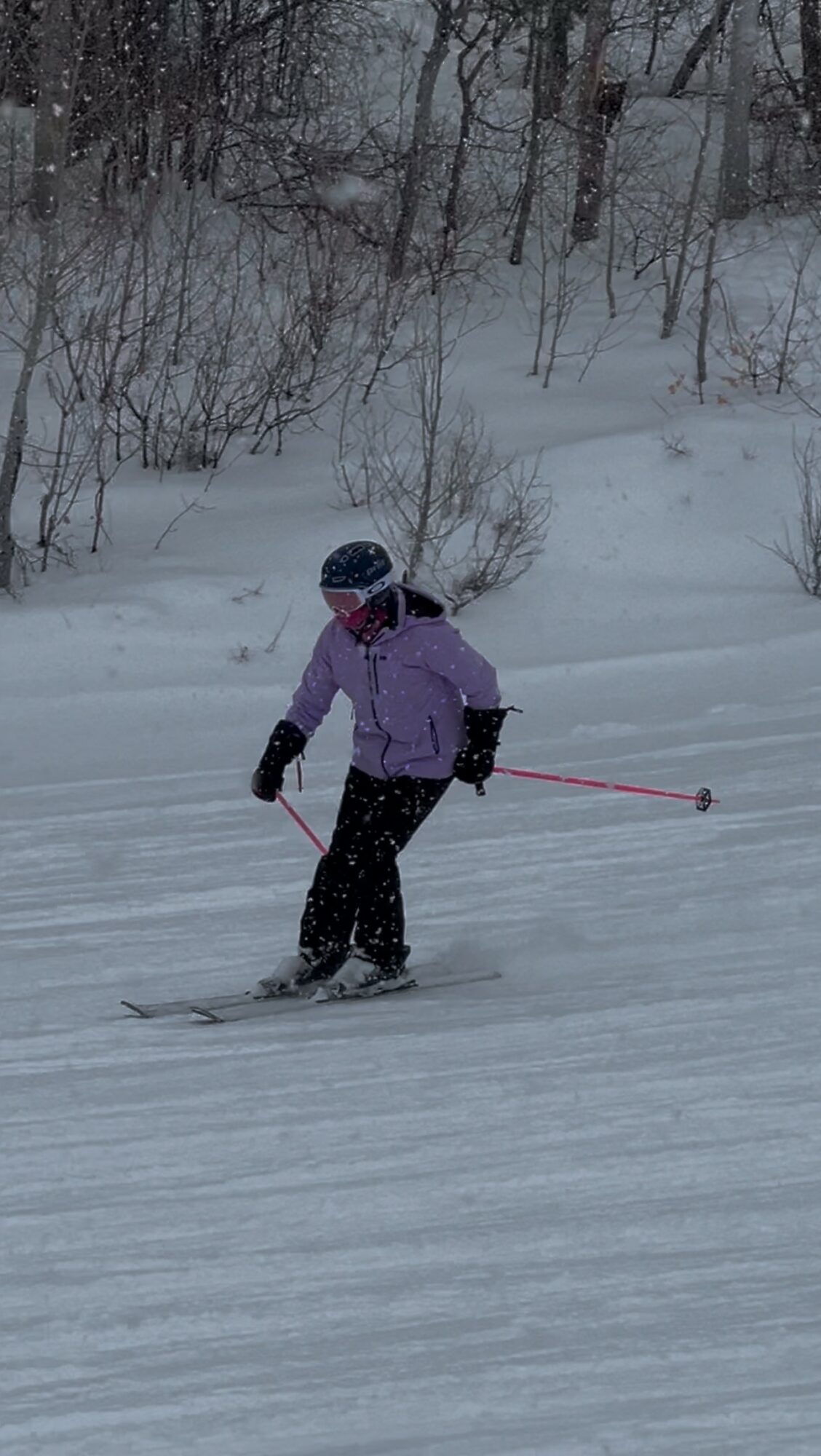 Child skiing on snow-covered ground with trees in the background, wearing a purple jacket and black helmet.