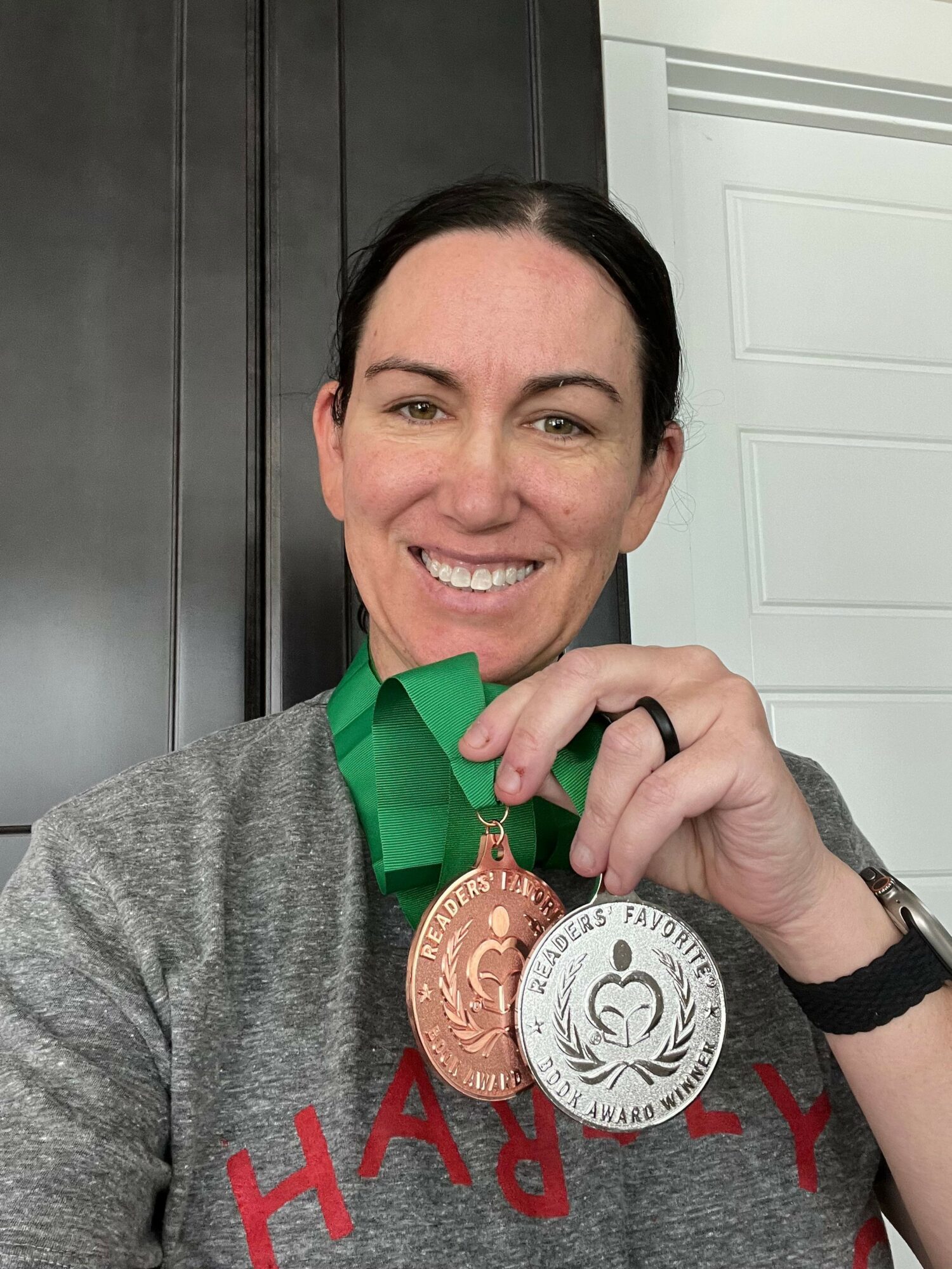 Woman smiling holding three medals, wearing a gray shirt with red text, in an indoor setting.