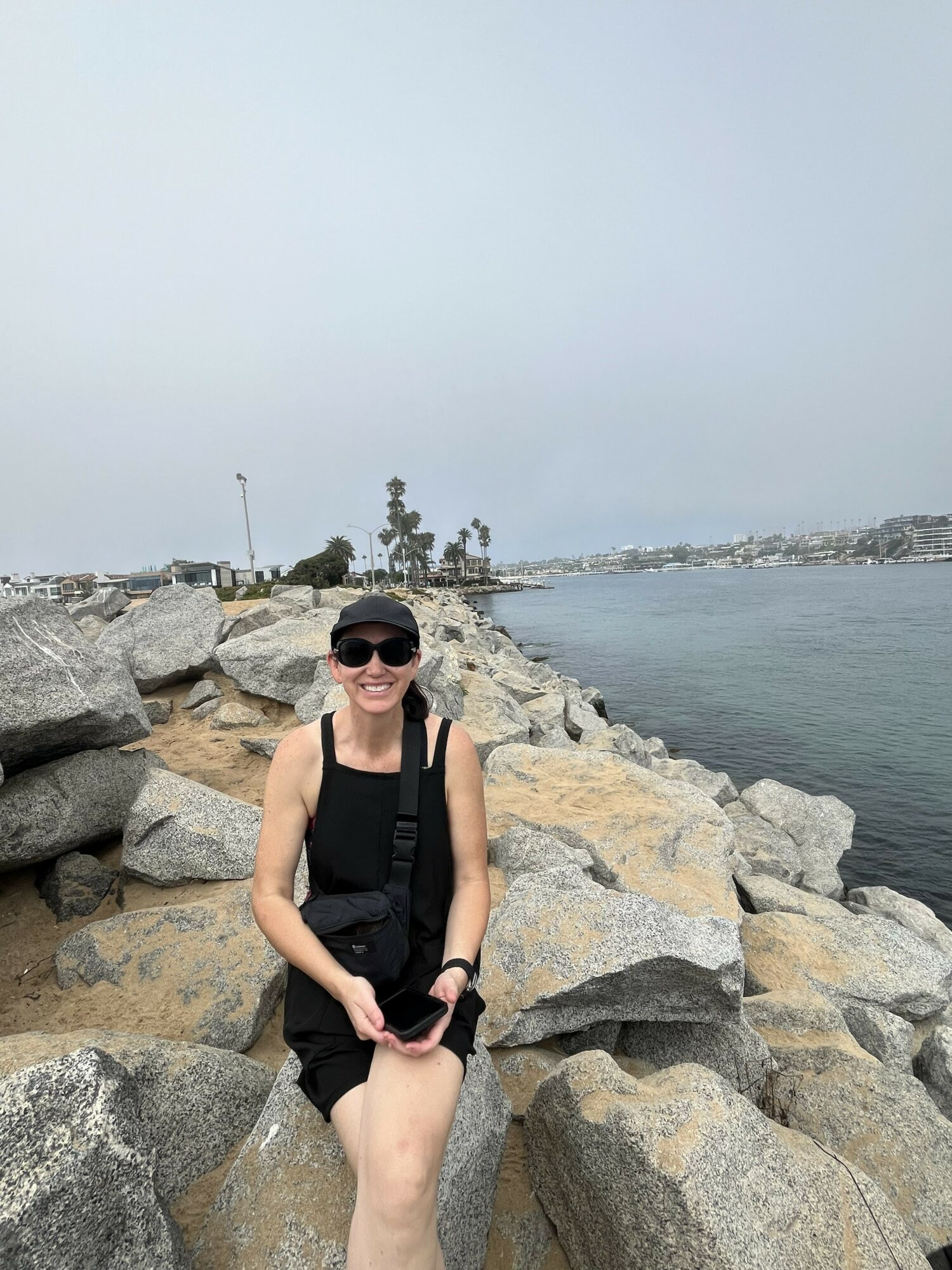 Woman in black outfit and sunglasses standing on rocks by water, smiling, holding a phone, overcast sky.