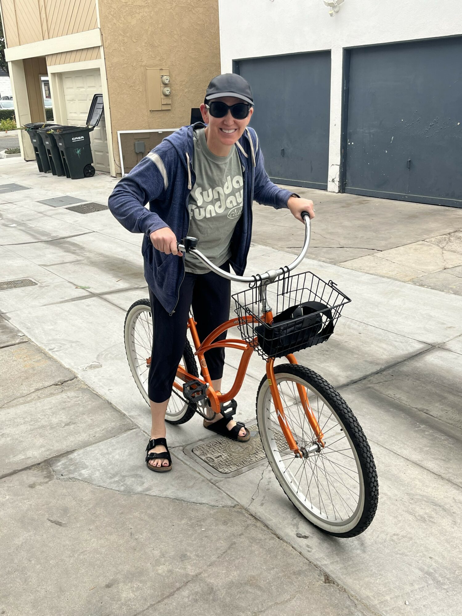 Person standing with an orange bicycle on a sidewalk, wearing sunglasses, a cap, and casual clothing, smiling at the camera.
