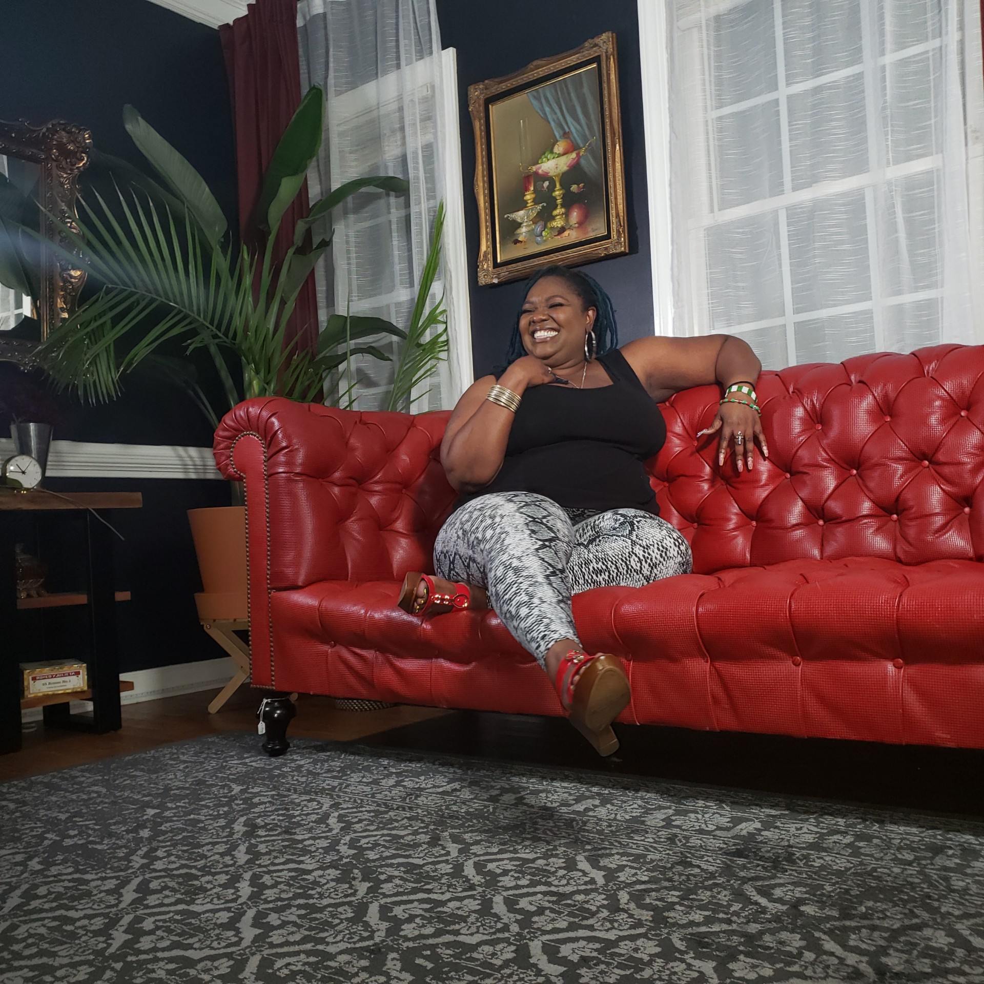 Woman sitting on a red sofa, smiling, in a room with large windows and plants.