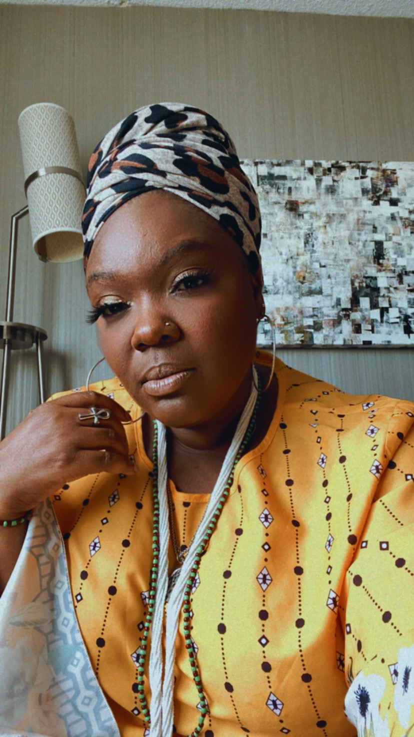 Woman wearing a patterned headscarf and yellow garment, sitting indoors with a textured wall and artwork behind her.