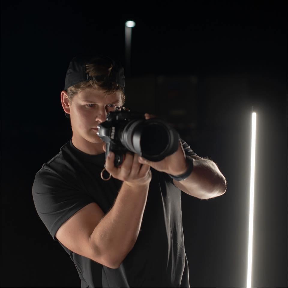 Young man holding a camera, taking a photo, with dark background and vertical light strip, wearing a black shirt and cap.