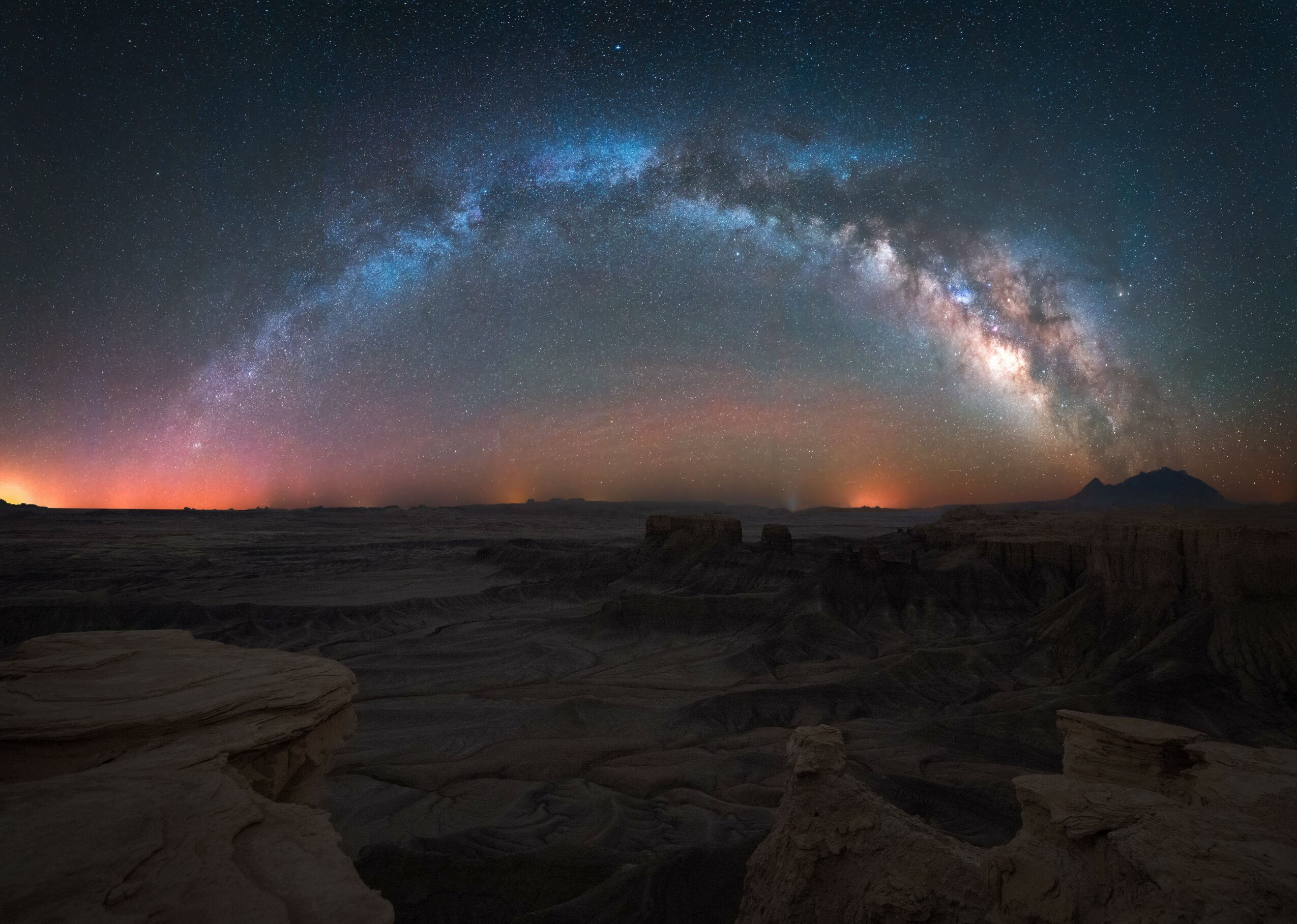 Night sky with a bright, arched Milky Way galaxy over a dark, rocky landscape.