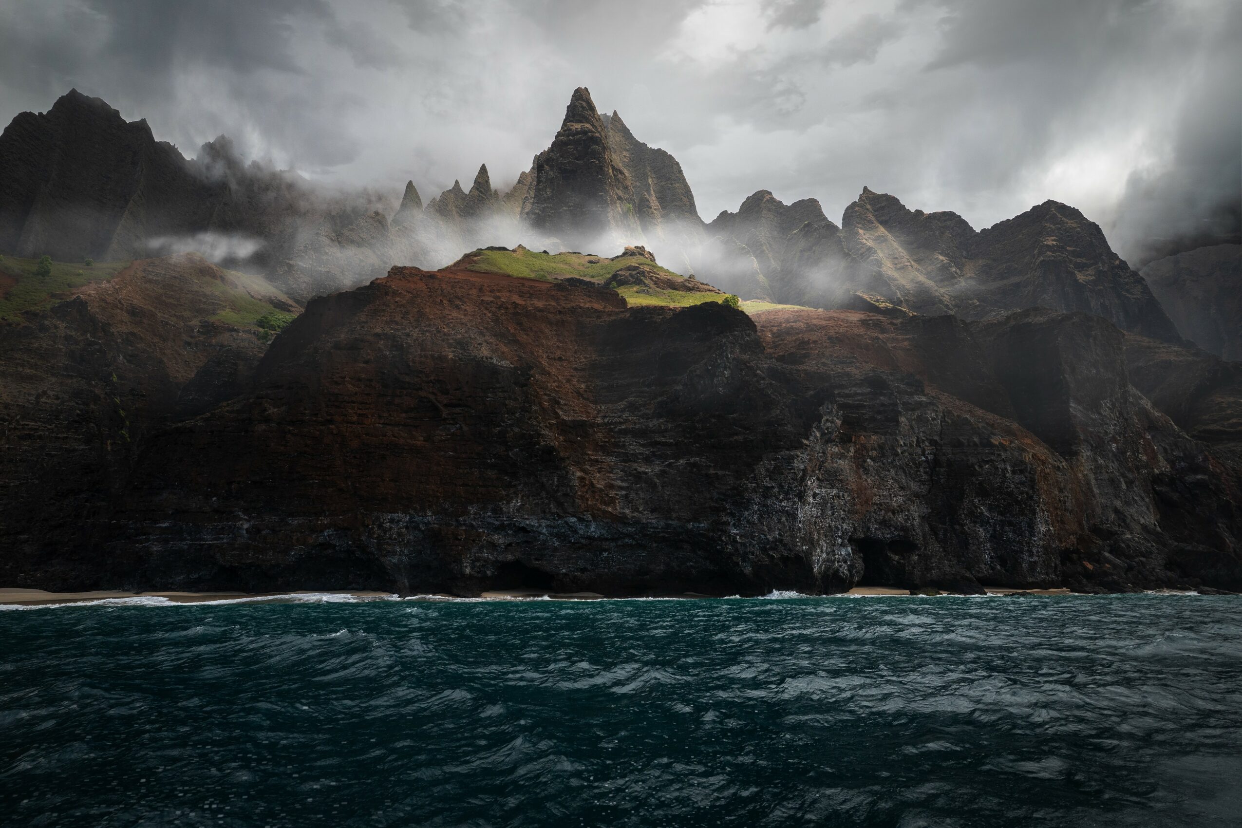 Cliffside with jagged mountain peaks under cloudy sky, ocean in foreground, mist around peaks, dark and moody atmosphere.