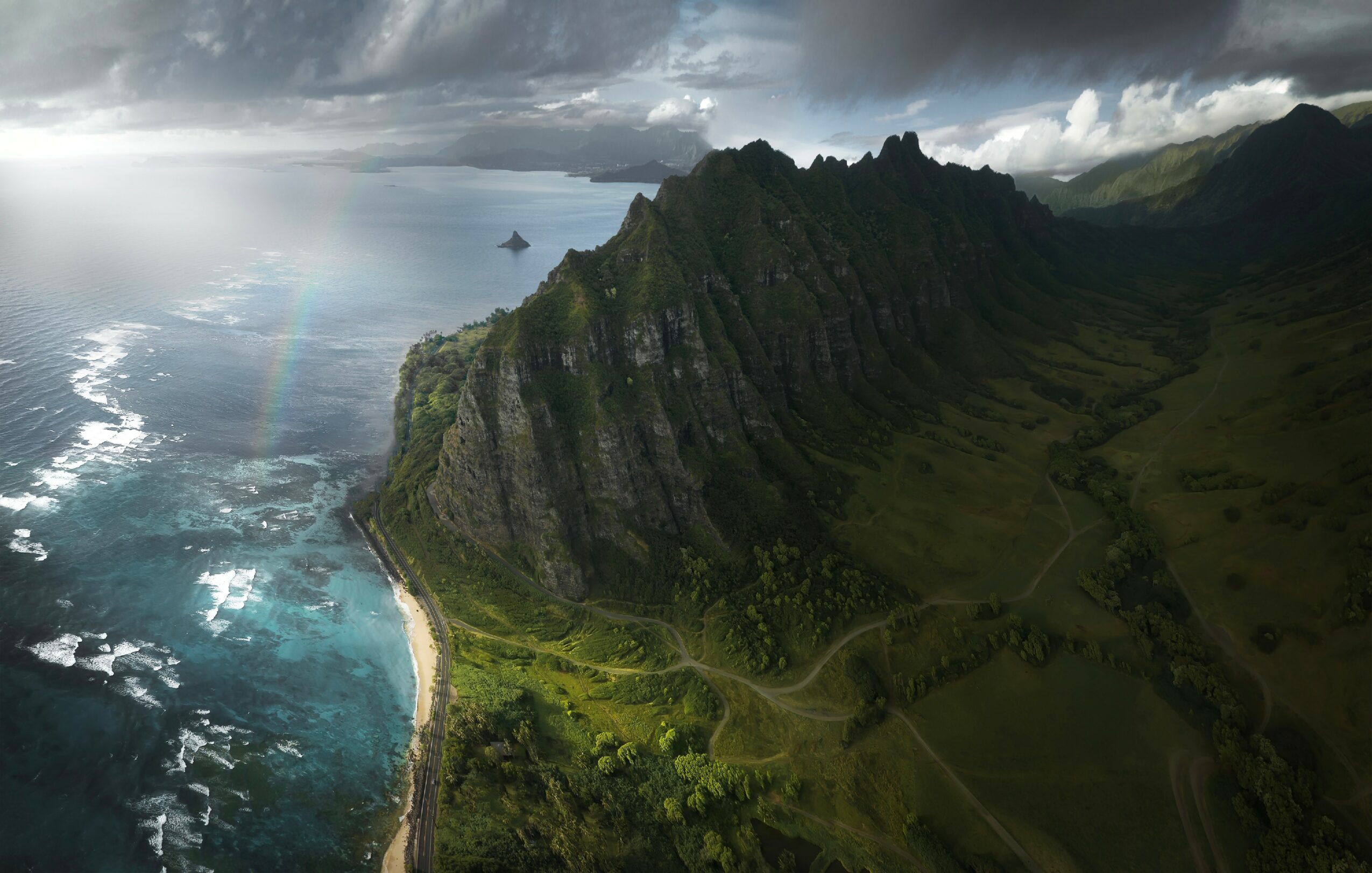 Cliffside landscape with steep green mountains, ocean, and cloudy sky, viewed from above.