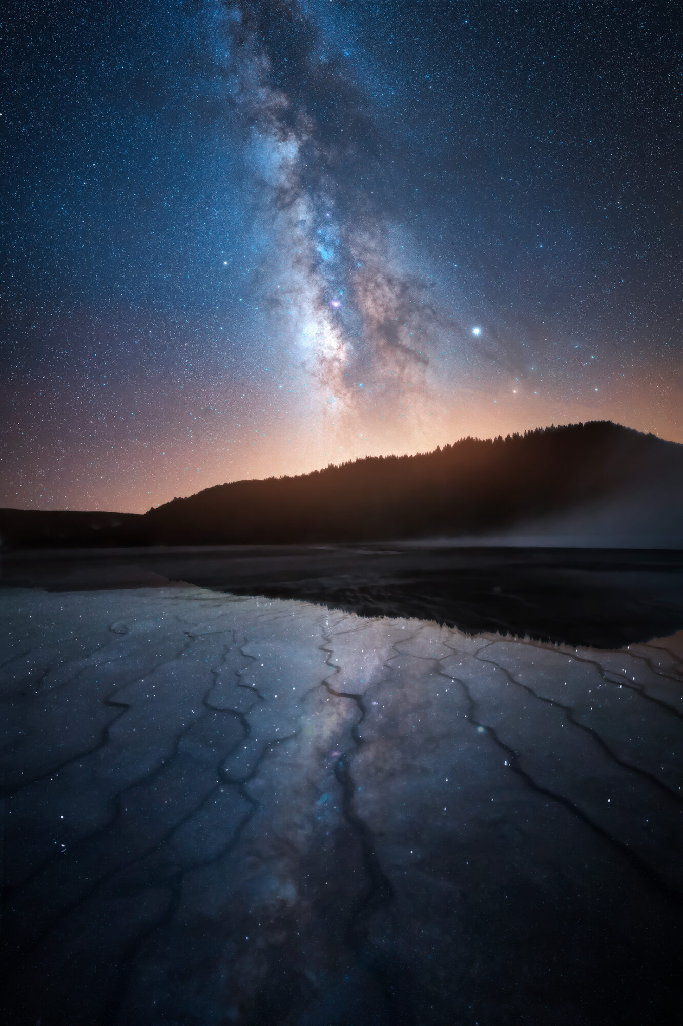 Night sky with the Milky Way galaxy above a dark landscape, reflected in a body of water, with cracked ground in foreground.