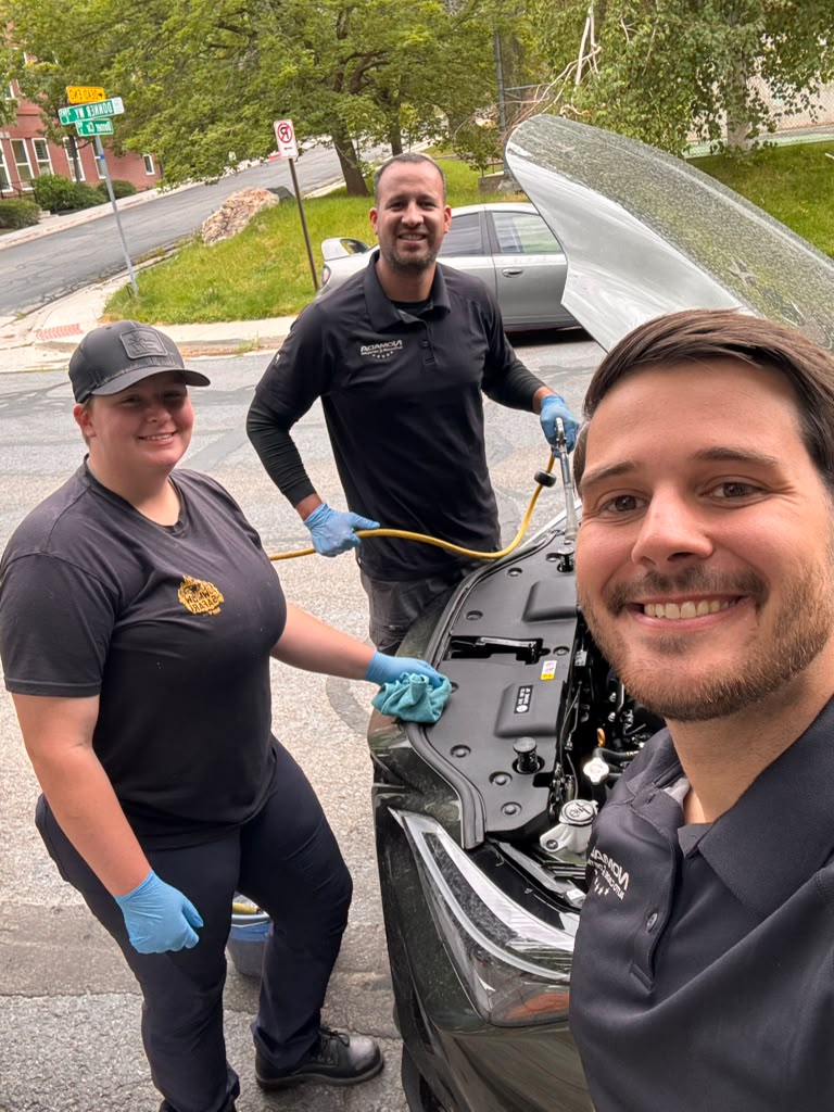 Three people working on a car engine outdoors, smiling, with trees and a street in the background.