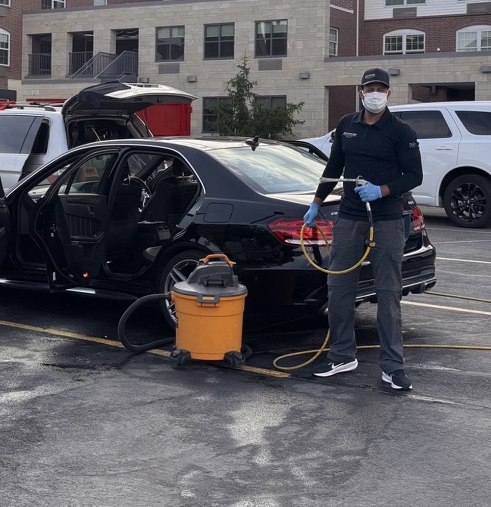 Person wearing a mask and gloves holding equipment next to a black car in a parking lot.