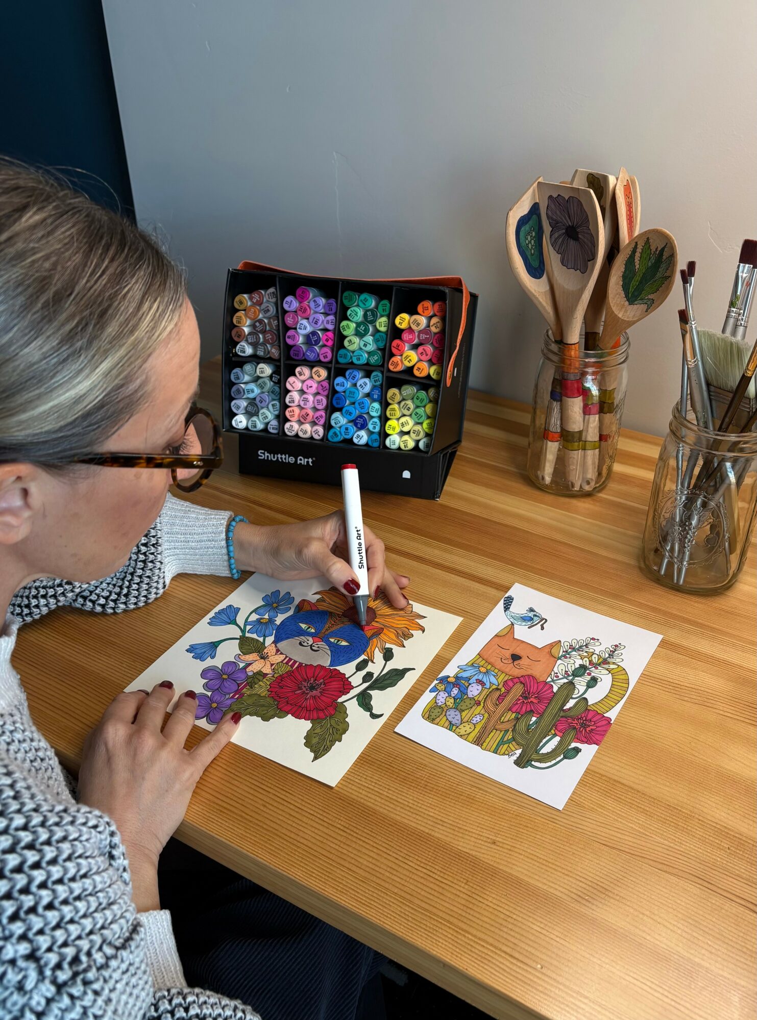 Person coloring floral and cactus illustrations with markers on a wooden table, with art supplies nearby.