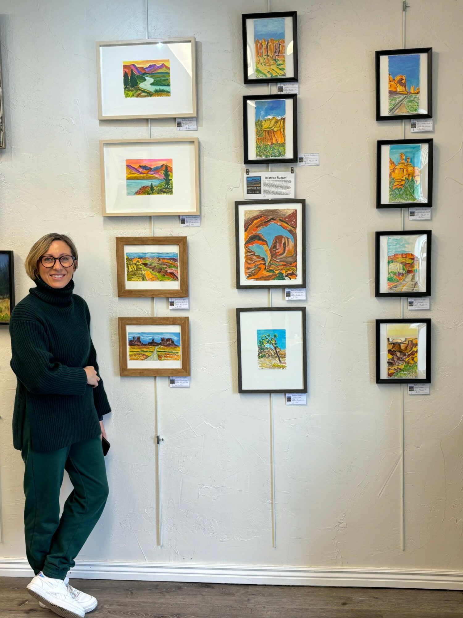 Person lying on the floor in front of a wall with framed artwork, smiling, wearing dark clothing and white shoes.