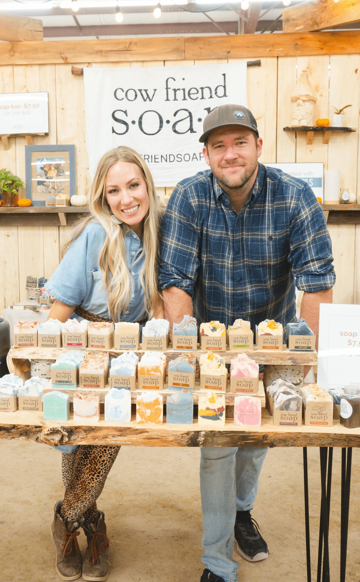 Two people, a woman and a man, stand behind a table with colorful soap bars, smiling at the camera, in a shop setting.