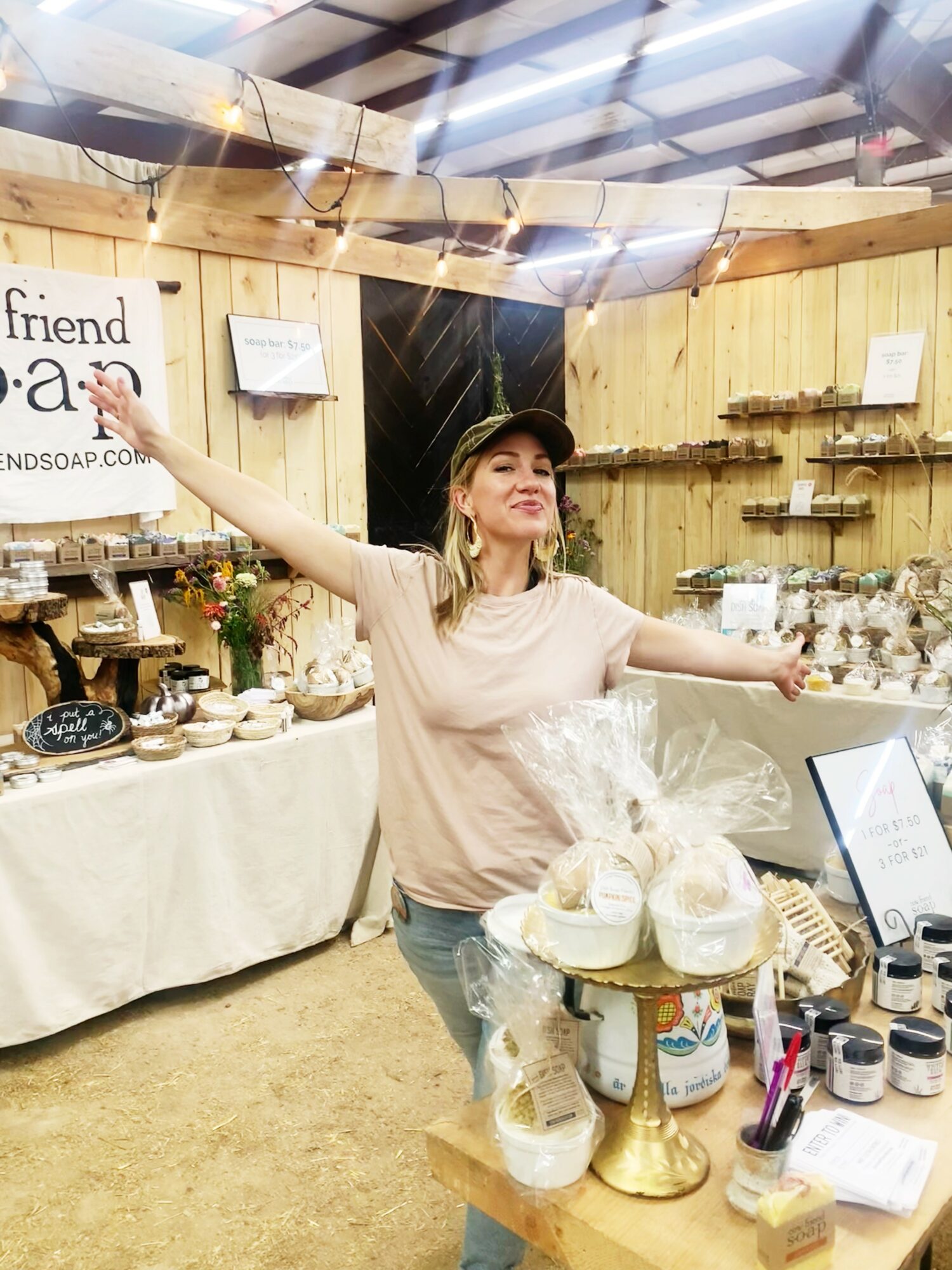 Woman with arms outstretched standing in a booth with wooden walls, displaying various items on tables and shelves.