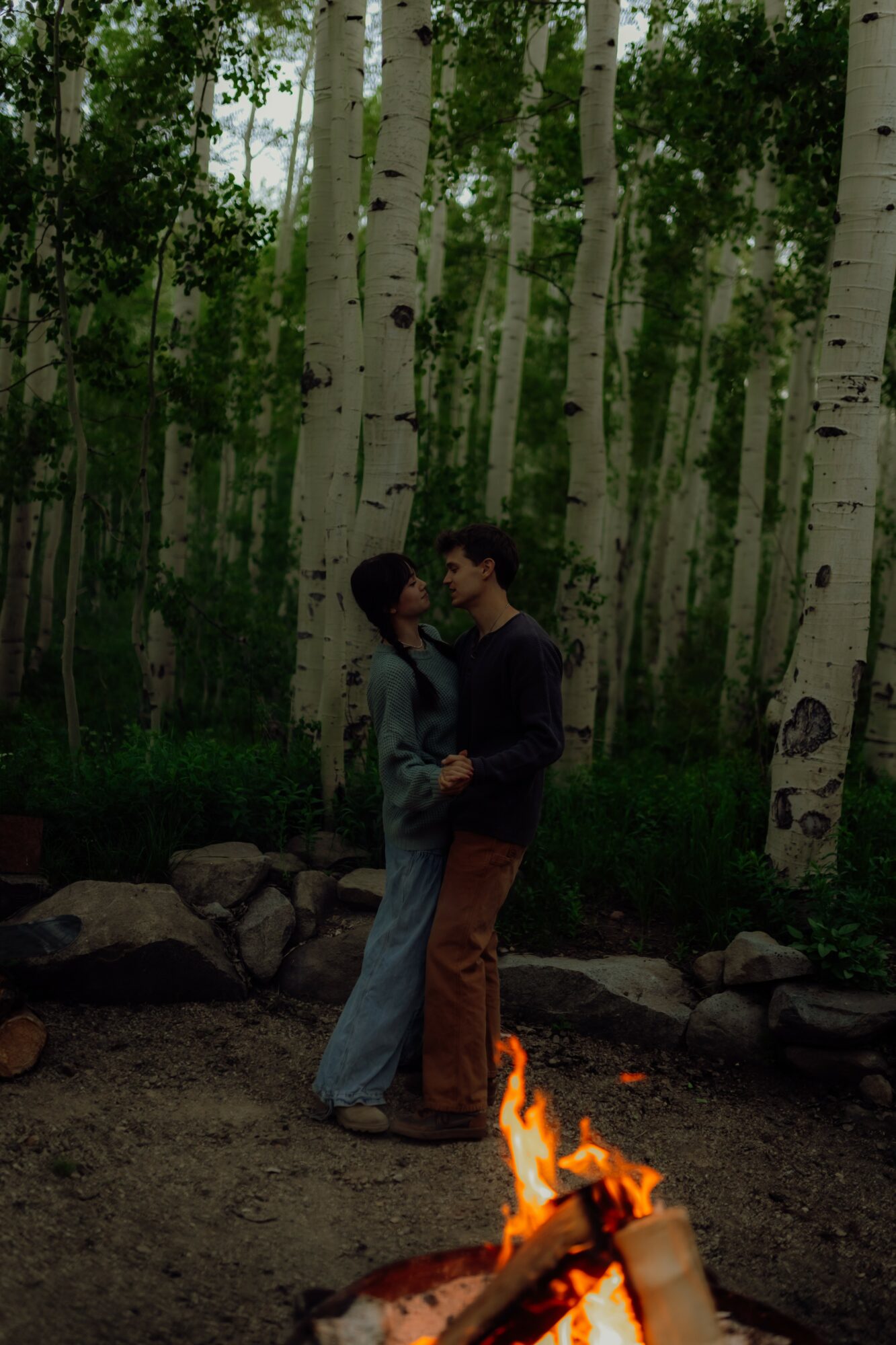 Two people stand close together near a campfire in a forest with tall white-barked trees.