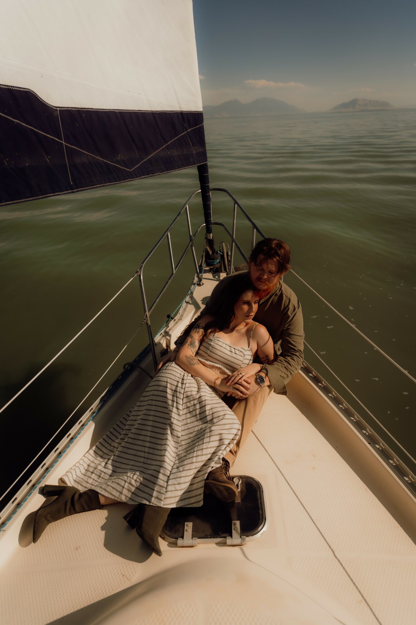 Two people sit on a sailboat's deck, with water and distant mountains in the background, under a clear sky.