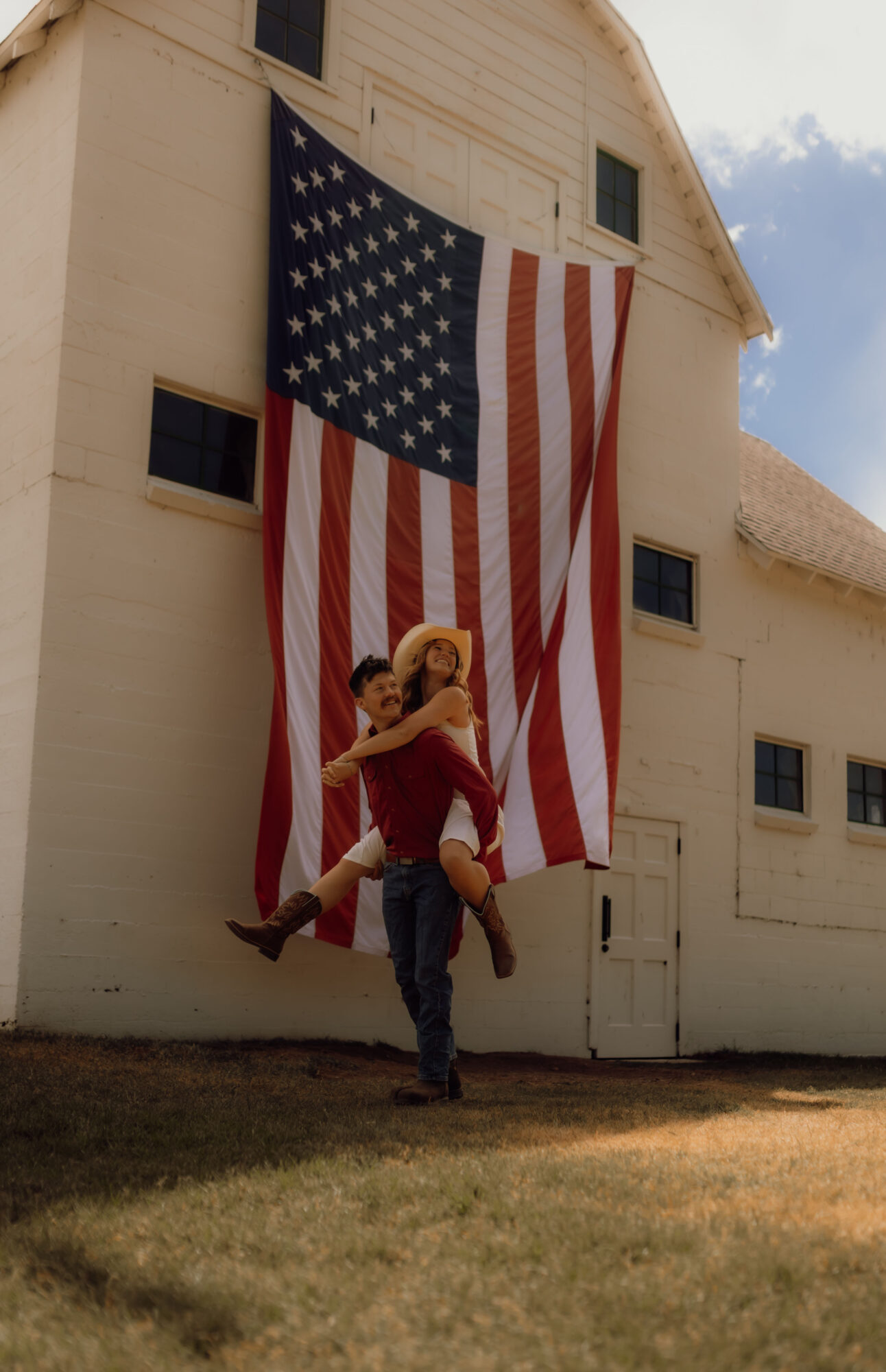 Person lifting child in front of a large American flag hanging on a white building, blue sky in background.