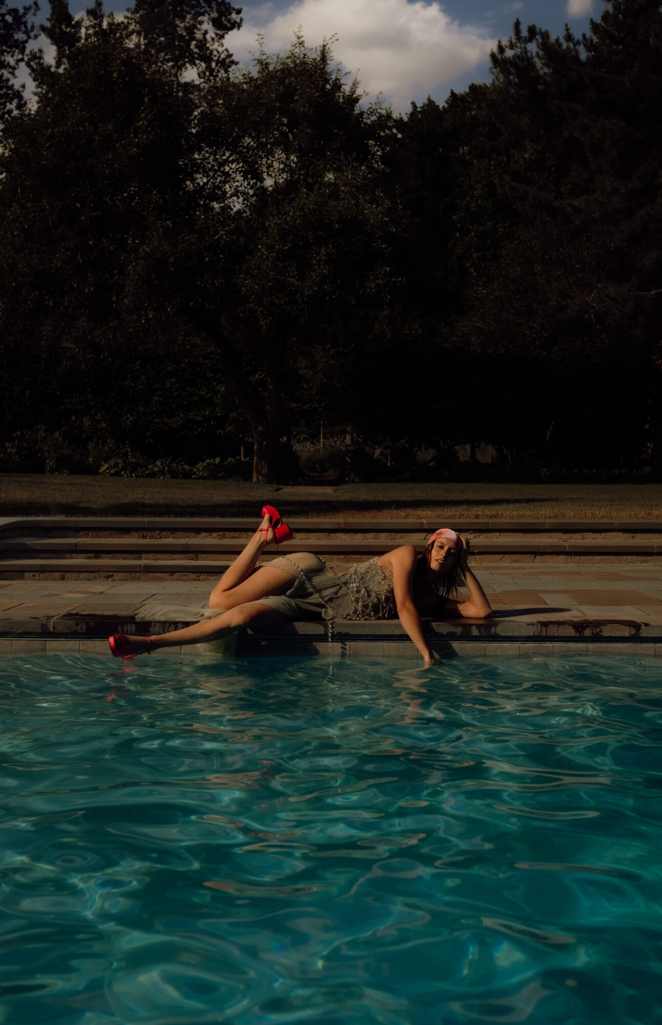 Two people at the edge of a swimming pool, one lying down and the other sitting, with trees and sky in the background.