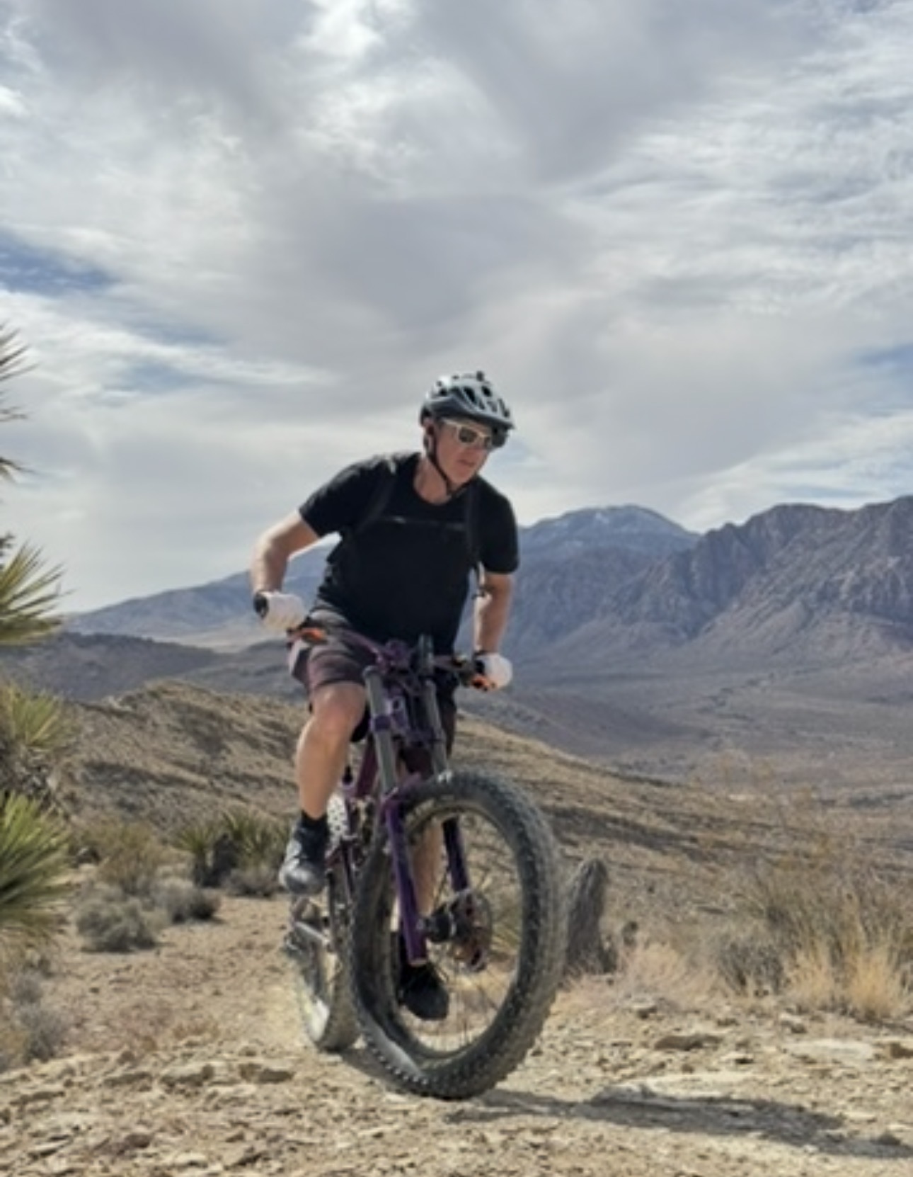 Person riding a mountain bike on a dirt trail in a desert landscape with mountains in the background, wearing a helmet and sunglasses.