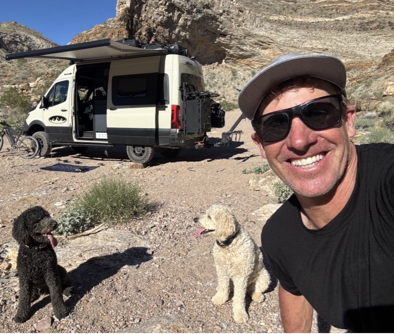 Man wearing sunglasses and a hat smiling outdoors with two dogs, a van, and rocky landscape in background.