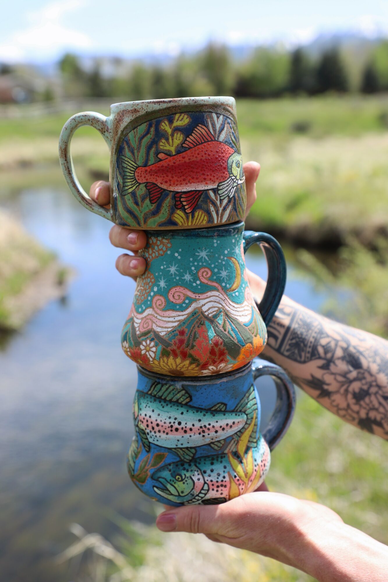 Four stacked ceramic mugs with colorful aquatic designs, held outdoors near a river with trees and sky in background.